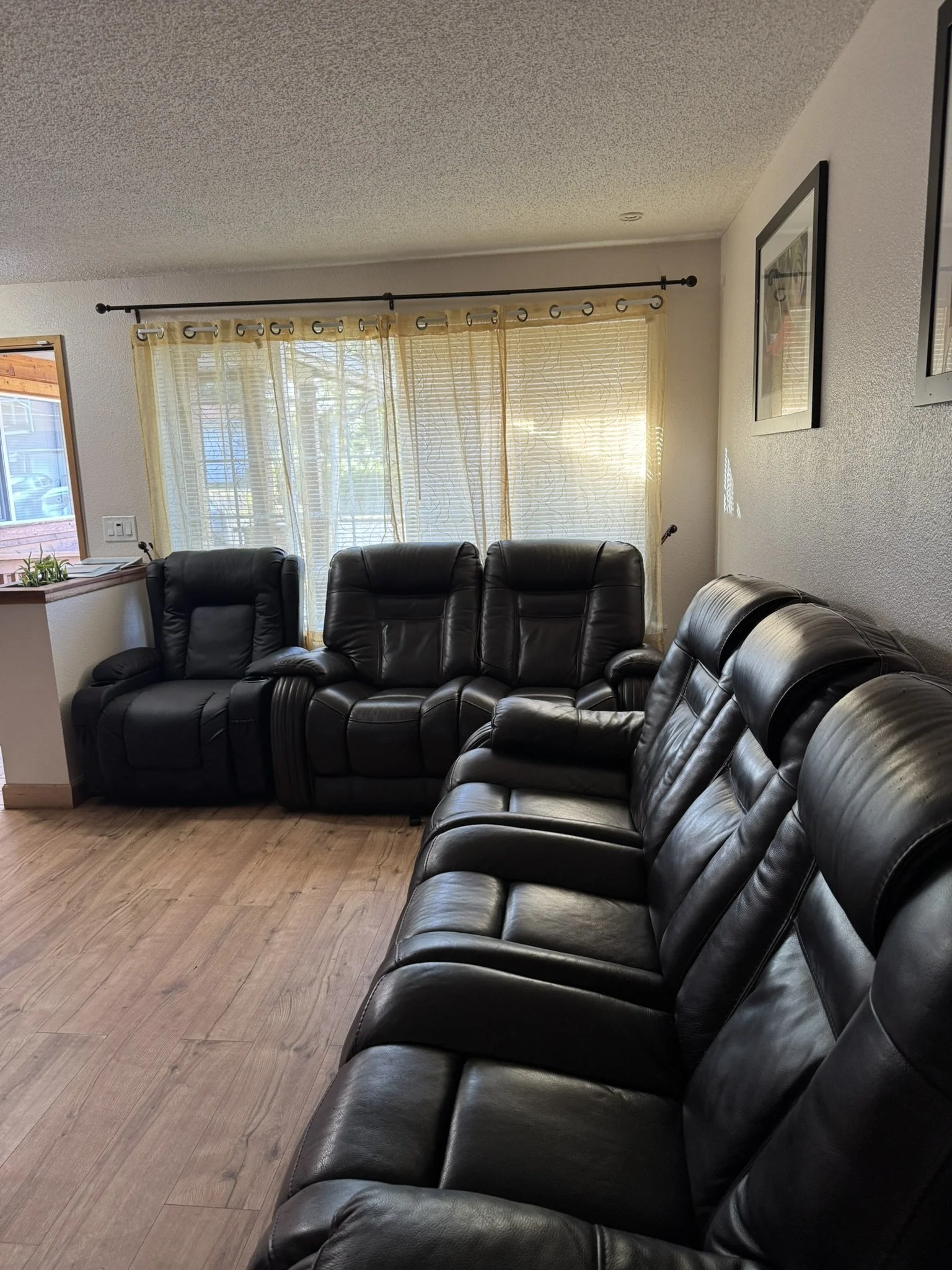 Living room with black leather recliner and sofa, yellow curtains, wooden floor, framed pictures on the wall, and city view outside the window.