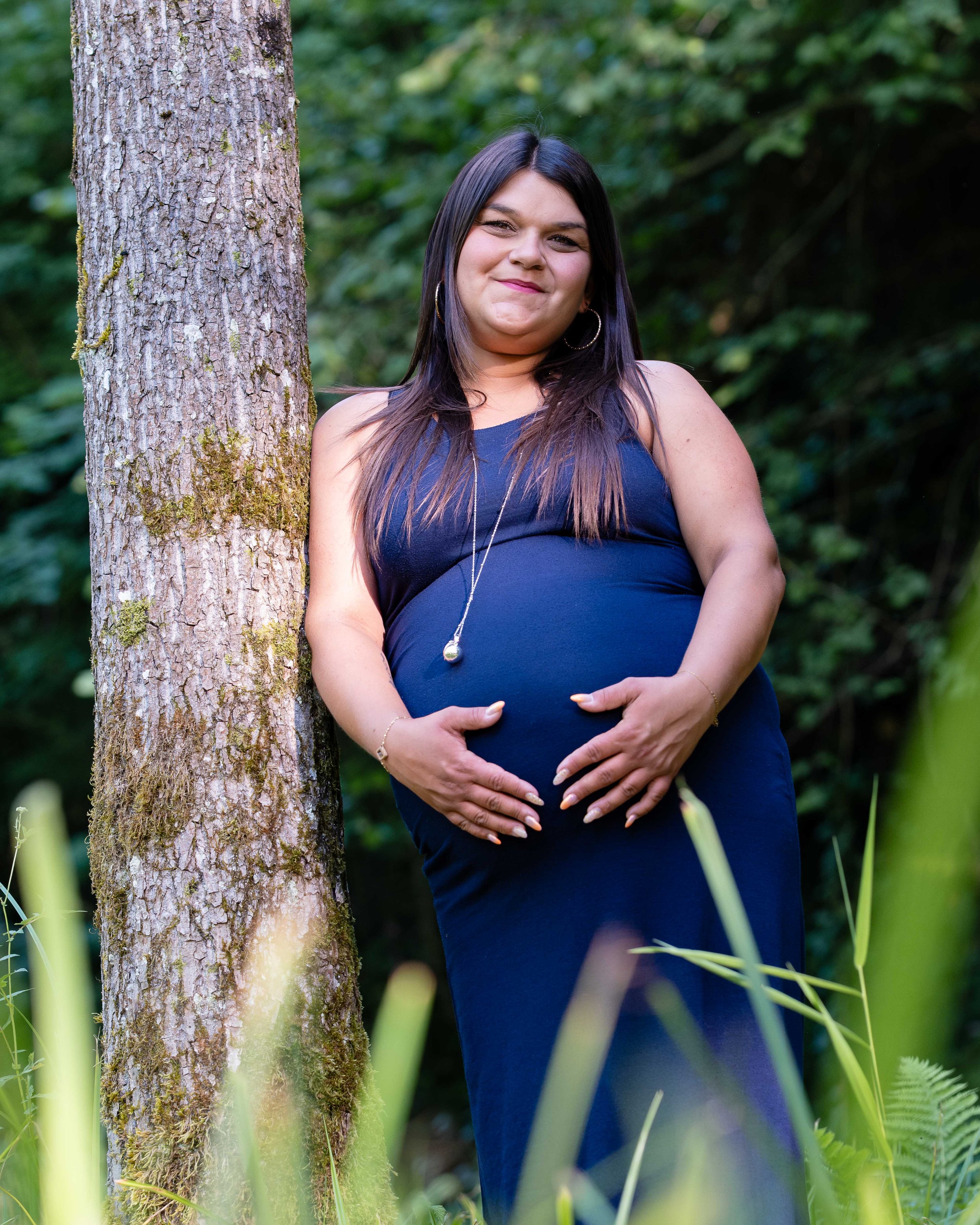A pregnant woman with dark hair and hoop earrings standing outdoors next to a tree with green foliage in the background. She is wearing a navy dress and is smiling while gently holding her belly with both hands.