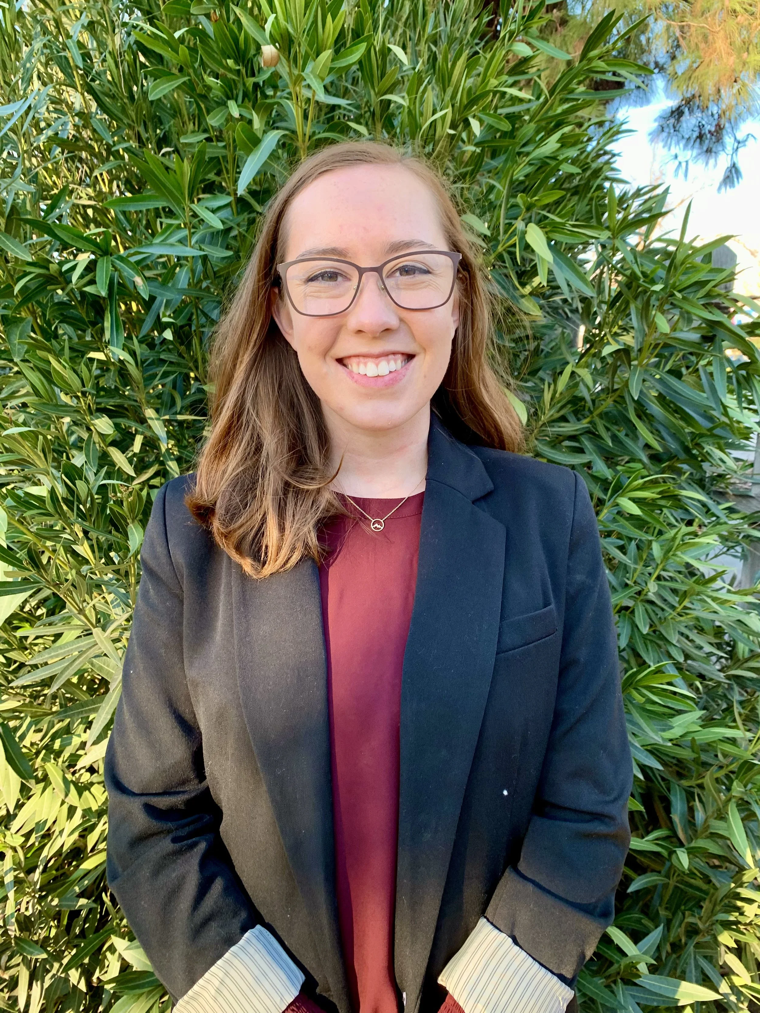 A smiling young woman with glasses, wearing a black blazer over a maroon shirt, standing outdoors in front of green foliage.