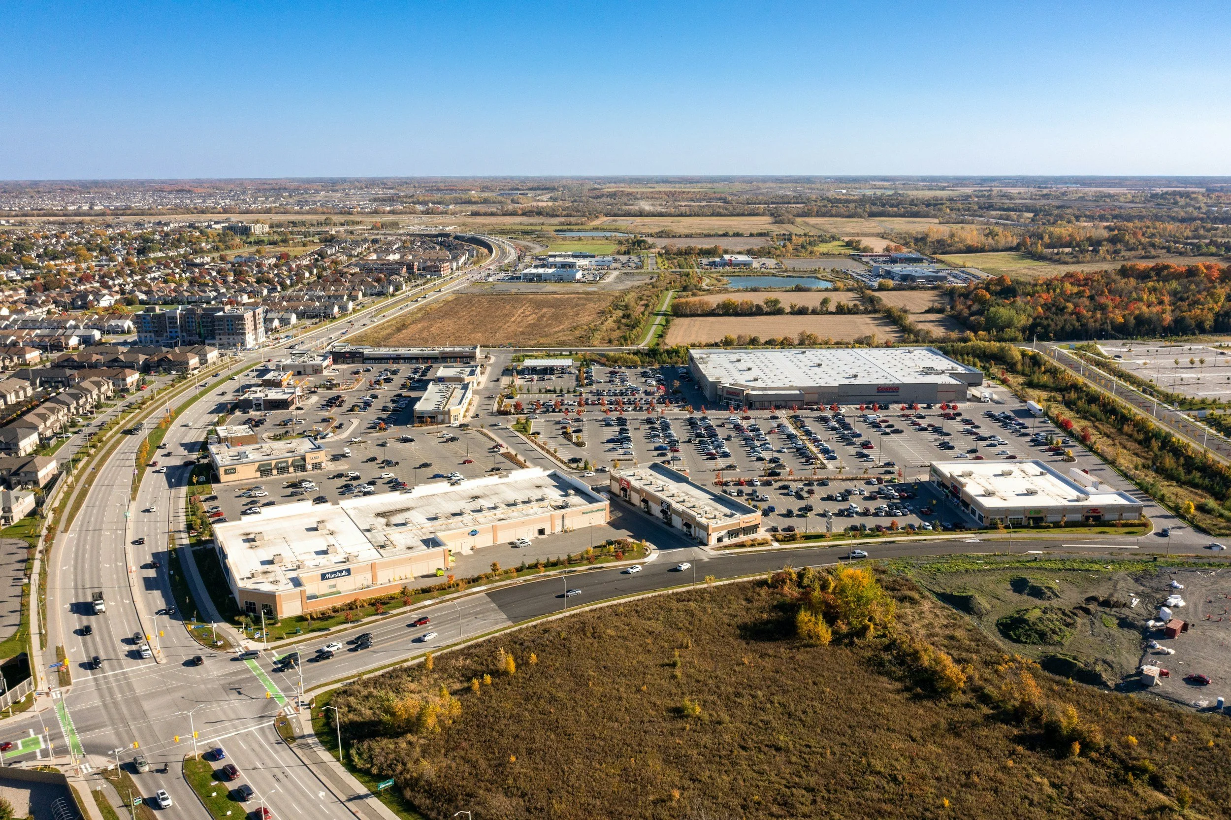An aerial view of a commercial shopping center with multiple stores, large parking lots, and surrounding roads, with residential neighborhoods and open fields in the background.