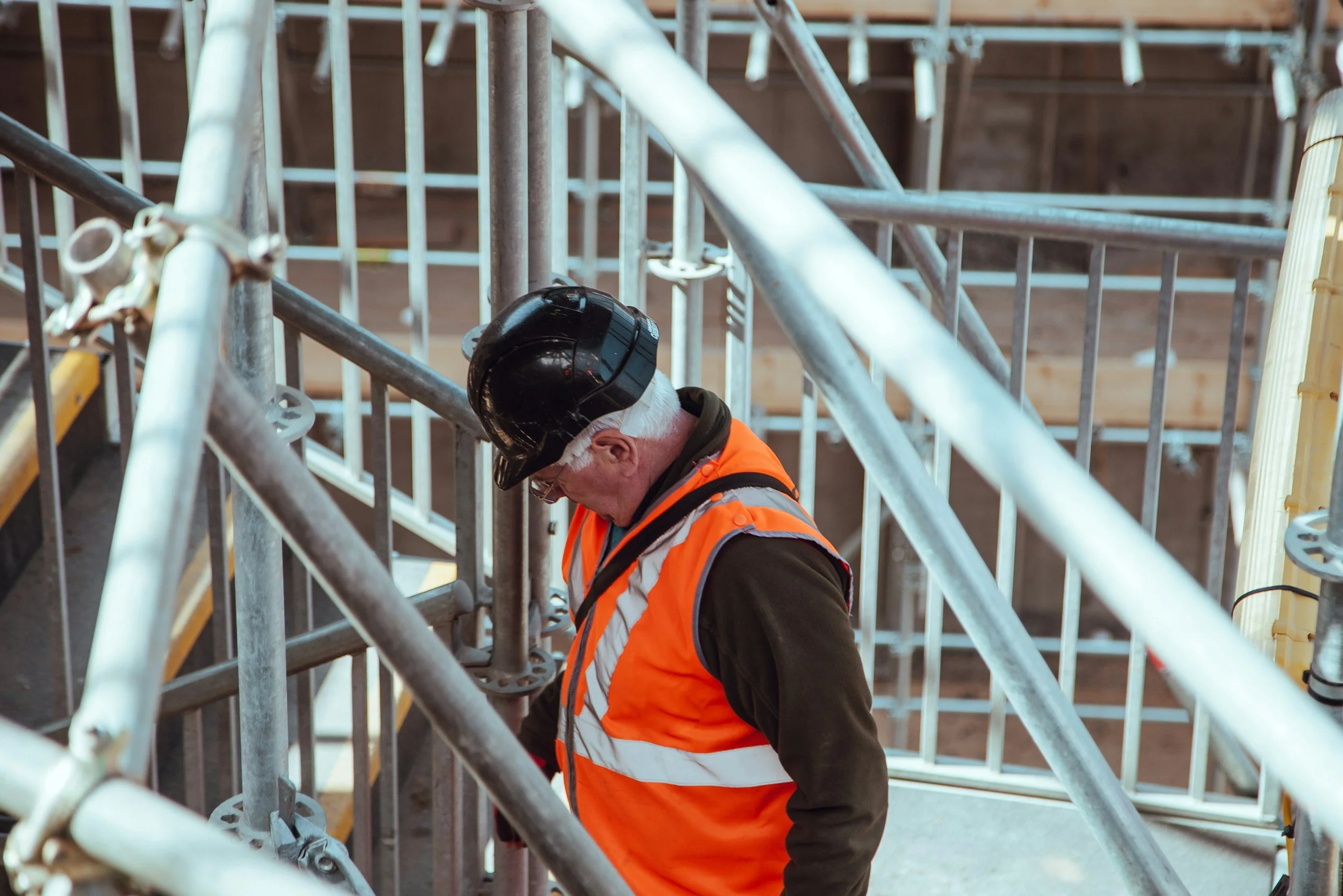 Construction worker in an orange safety vest and a black helmet working inside metal scaffolding at a construction site.