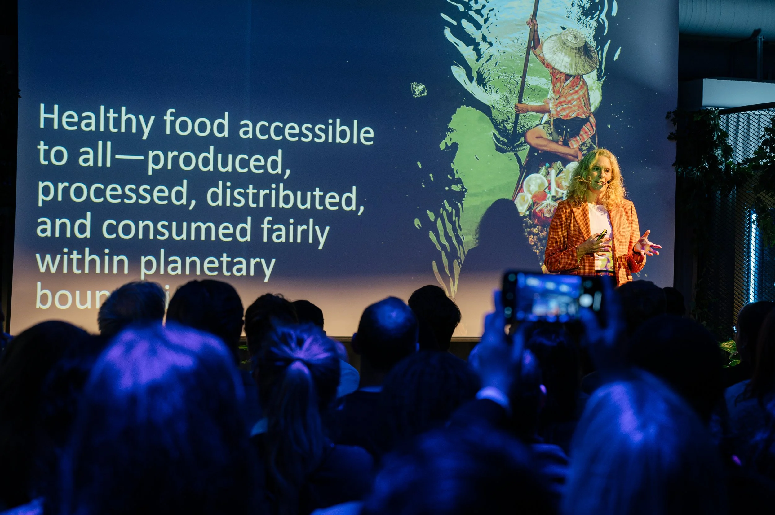 A woman presents on stage in front of a large screen displaying a slide about healthy food accessibility. The audience is seated and some are taking photos.