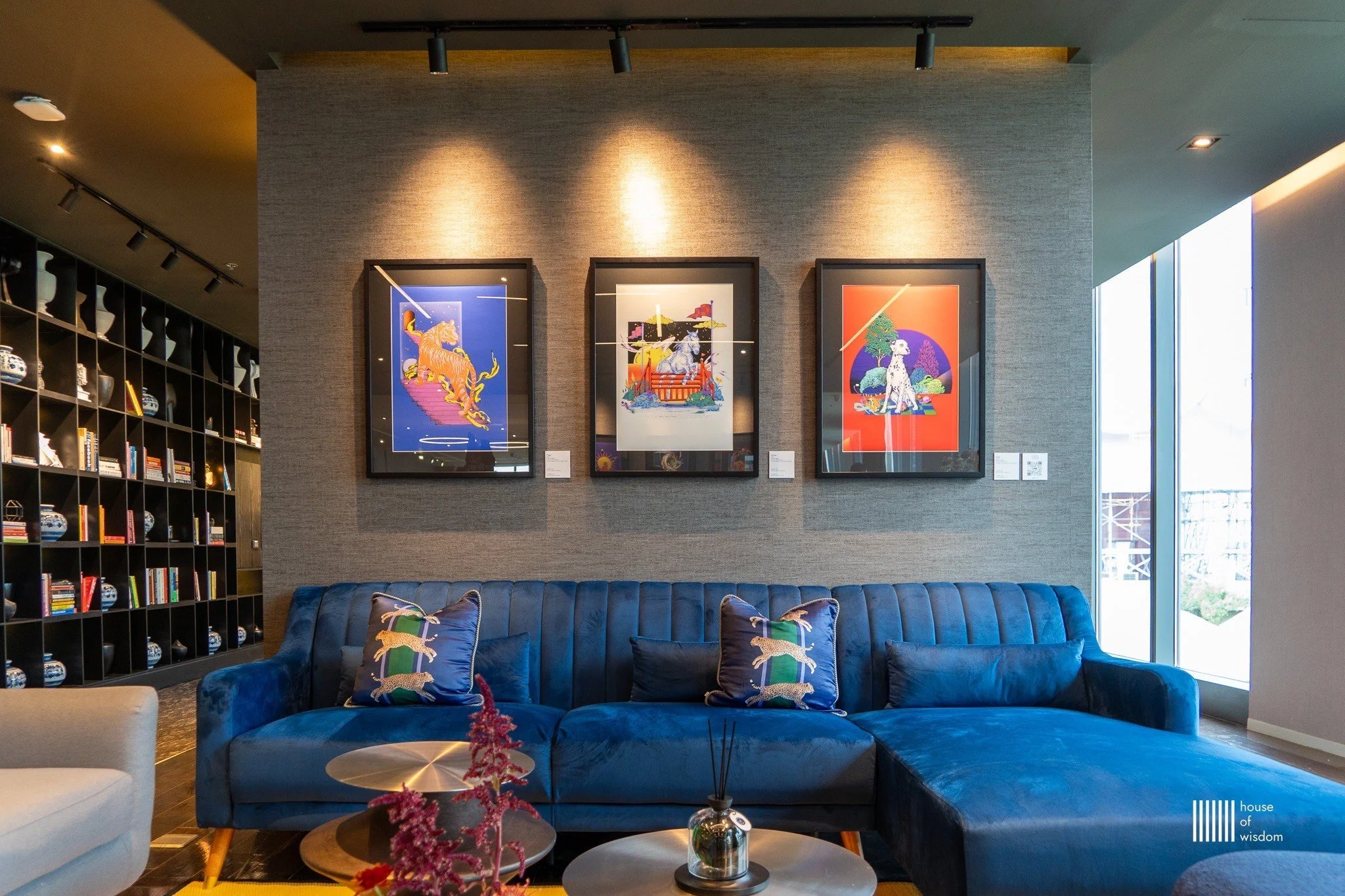 Modern lounge with blue velvet sectional sofa, decorative pillows, a bookshelf with ceramics and books, and three framed colorful art pieces on a textured wall.
