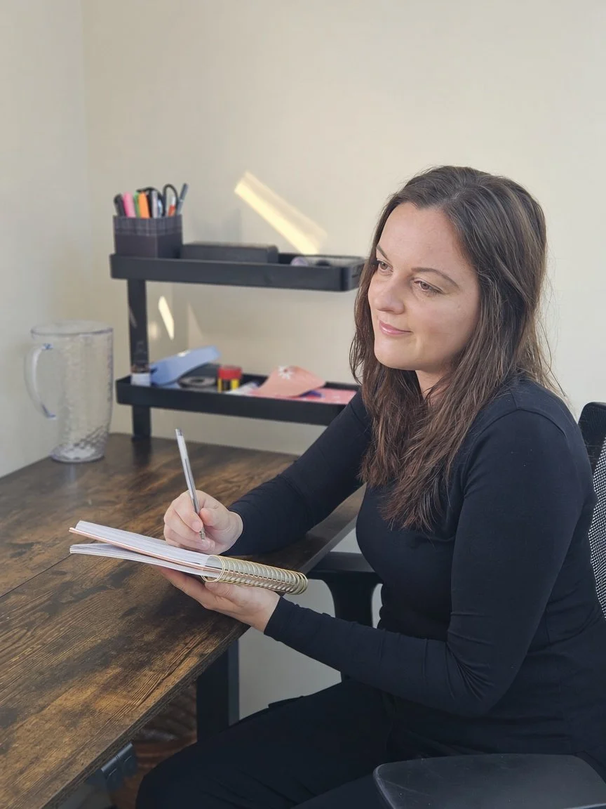 A woman with long brown hair sitting at a wooden desk, writing in a spiral notebook with a pen.