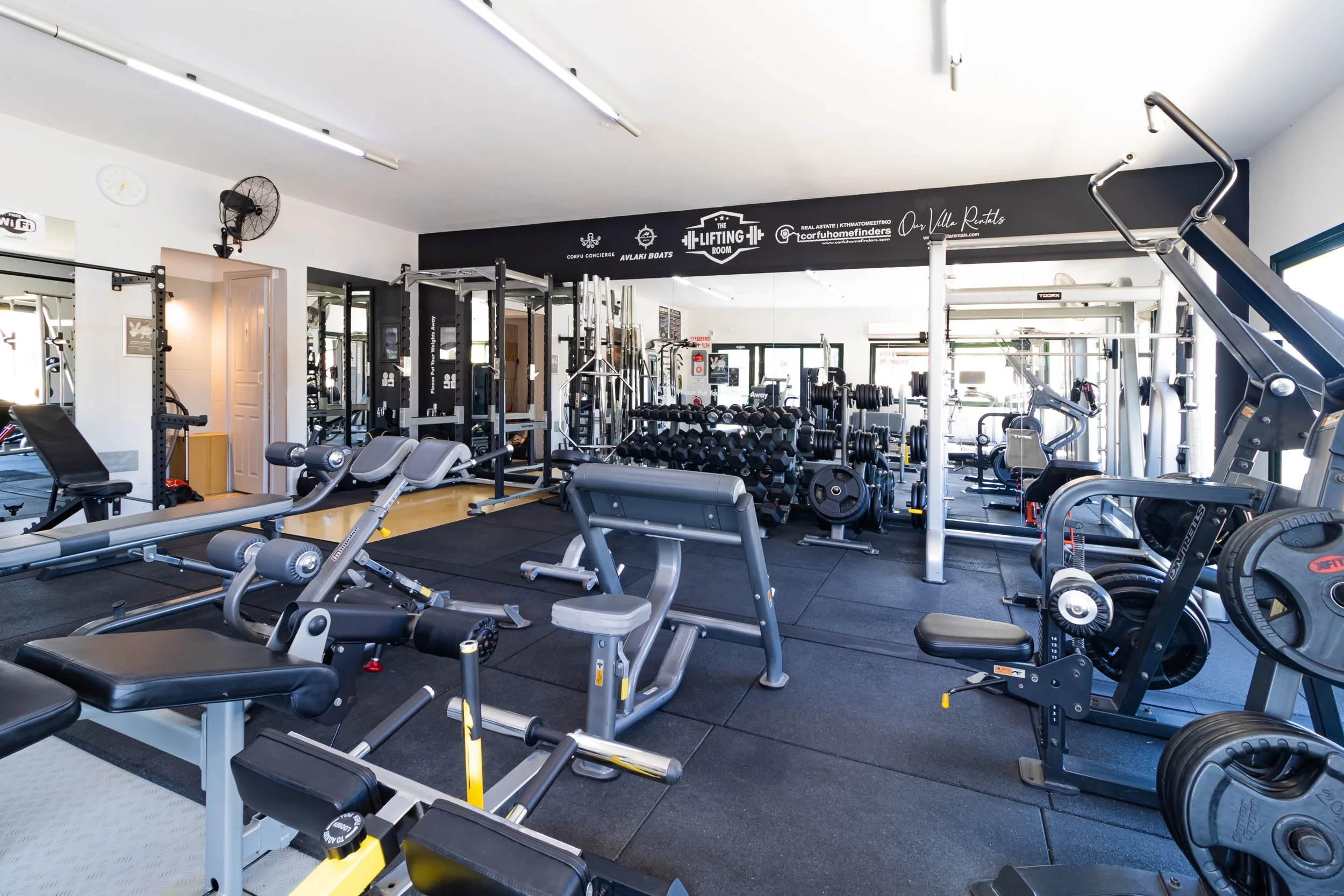 A modern gym interior with various fitness equipment including weights, benches, and exercise machines. There's a prominent sign reading "The Lifting Room" and mirrors on the walls. The space is well-lit, featuring a clean, organized layout.