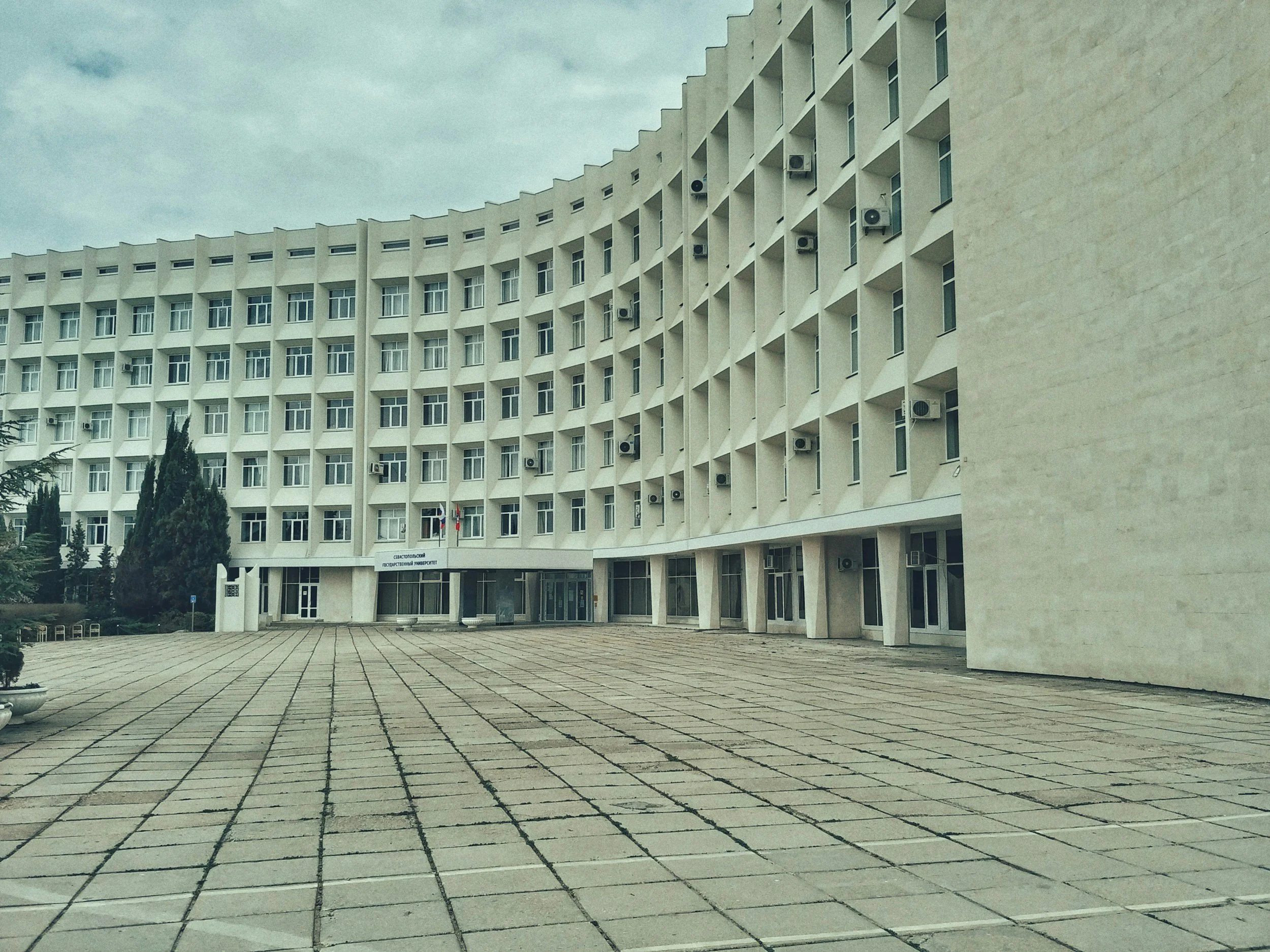 A large white multi-story building with numerous windows and air conditioning units, with a paved foreground and cloudy sky.