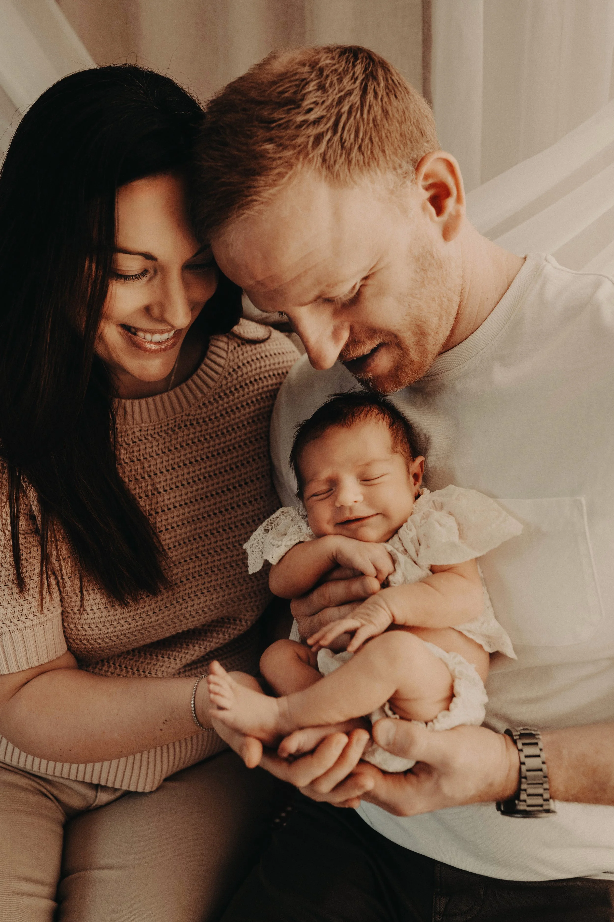 A woman, a man, and a baby are close together, smiling and looking at each other. The woman has long dark hair and is wearing a pink sweater. The man has short reddish hair and is wearing a gray shirt. The baby is being held by the man, wearing a light-colored outfit, smiling with eyes closed, with a loving family moment.