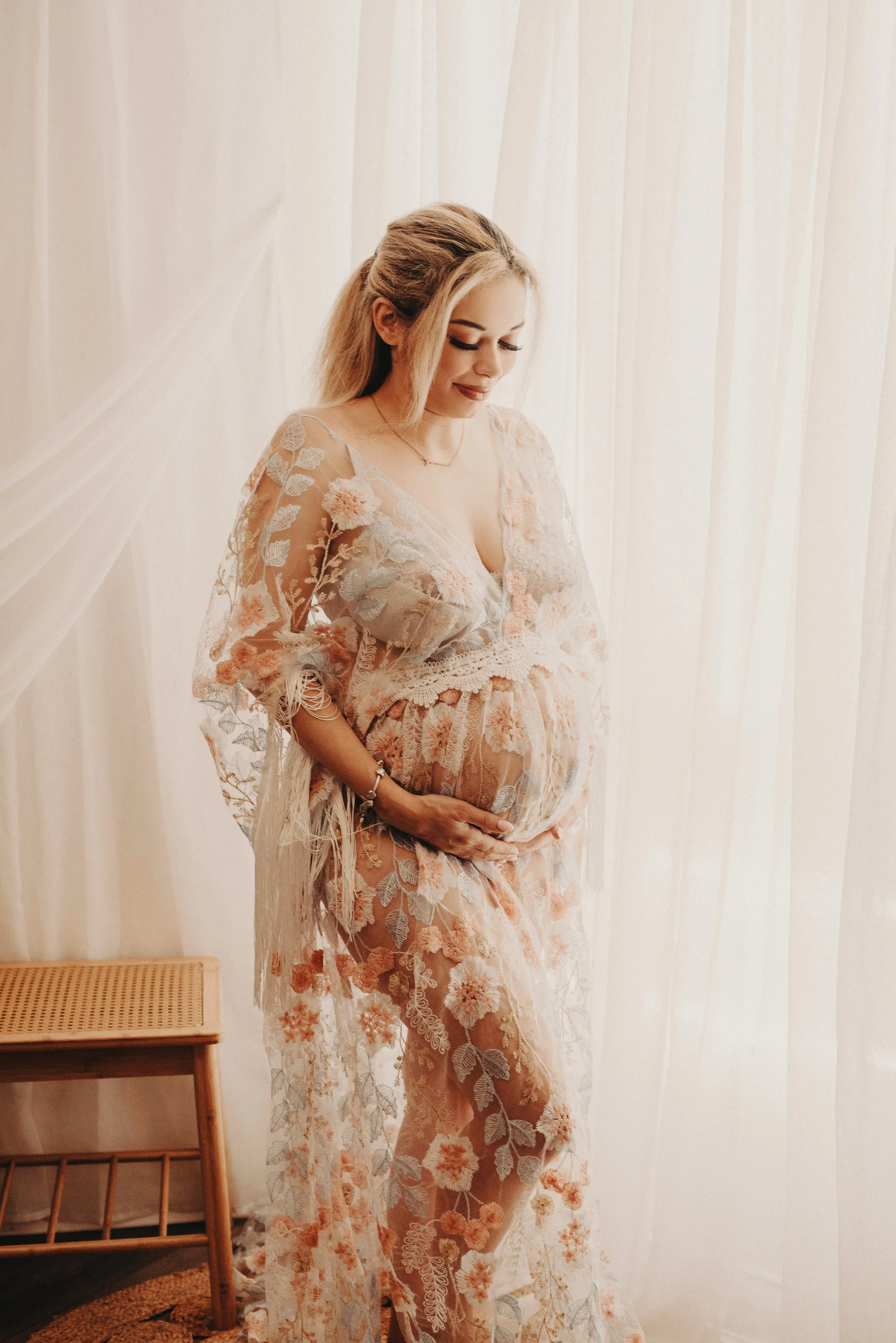 A pregnant woman with blonde hair in a loose style, wearing a sheer, embroidered dress with floral patterns in pastel shades, standing in front of a cream-colored curtain with a gentle smile and her hands resting on her belly.
