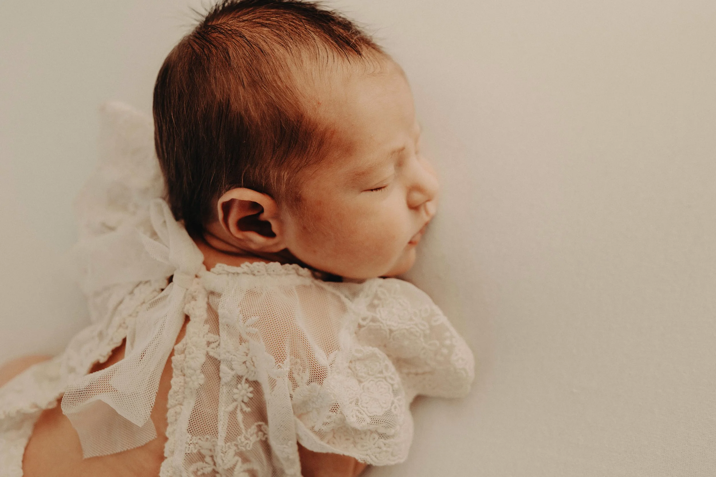 Close-up side view of a baby with closed eyes, lying against a plain, light-colored background, wearing a white lace outfit.