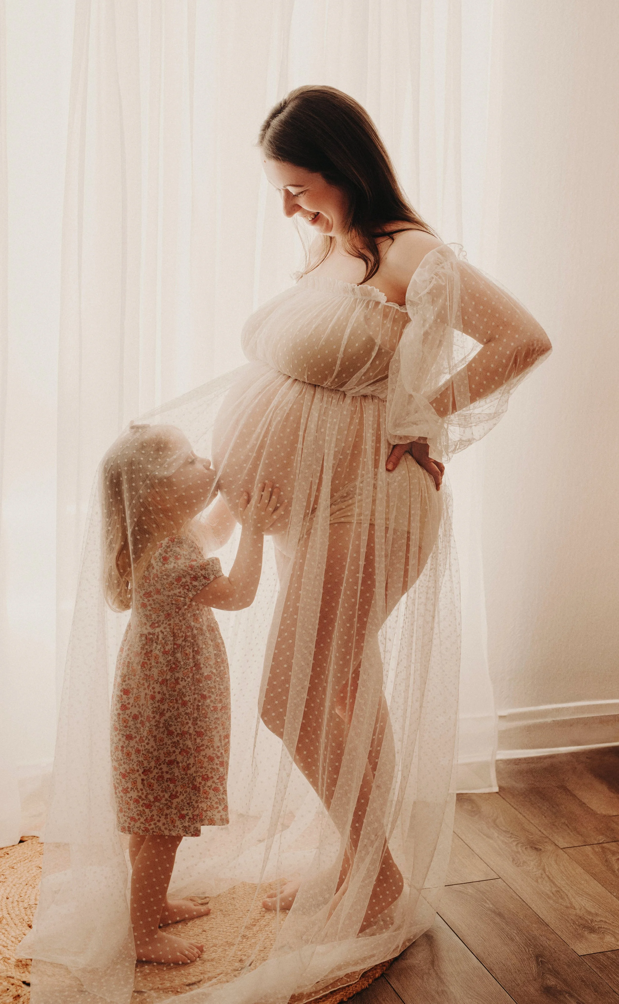 A pregnant woman and a young girl stand behind a sheer curtain, smiling and touching her belly, in a softly lit room.