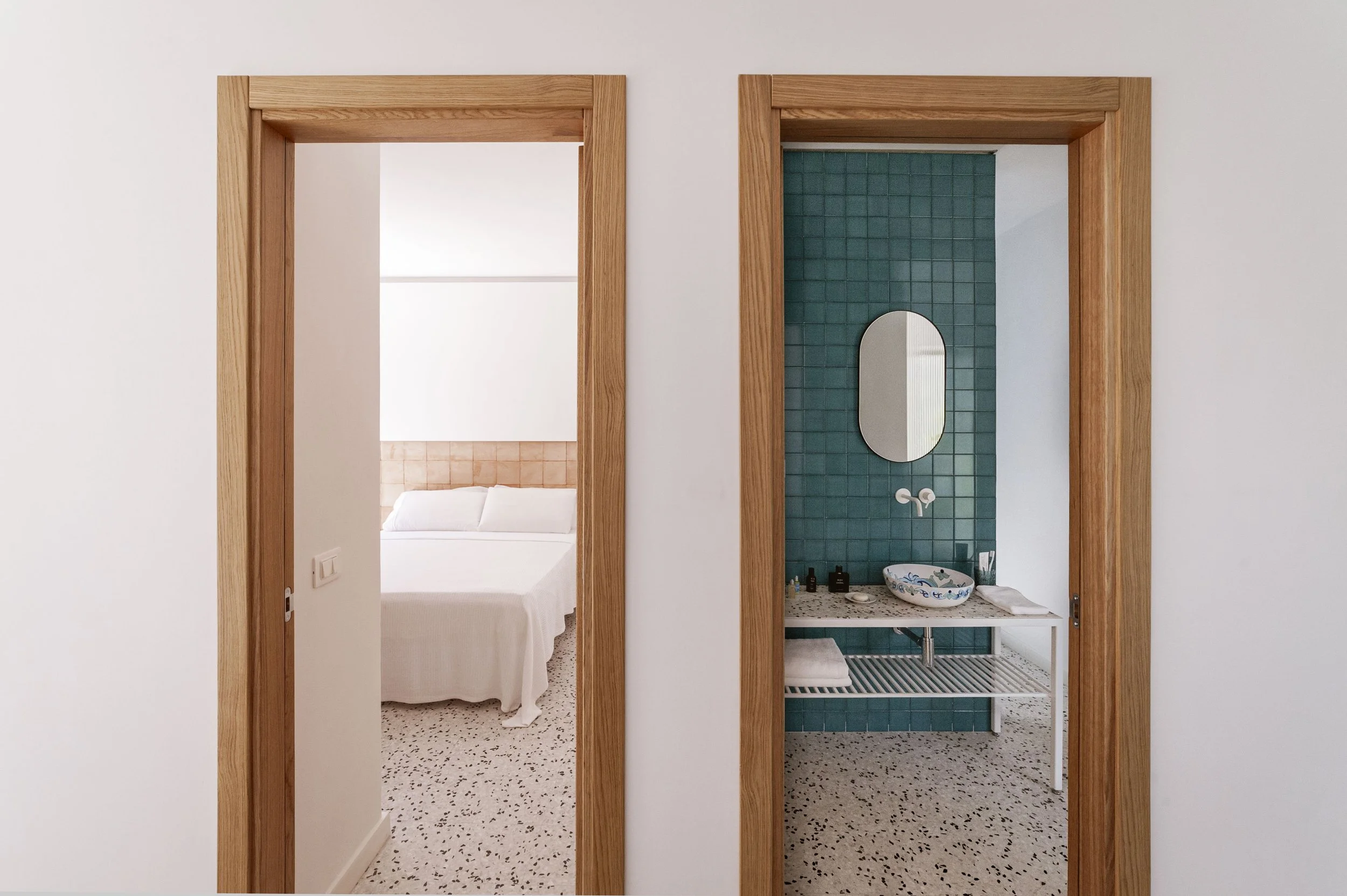 View of a bedroom with a white bed and headboard and an adjacent bathroom with a green tiled wall, mirror, sink, and toiletries.