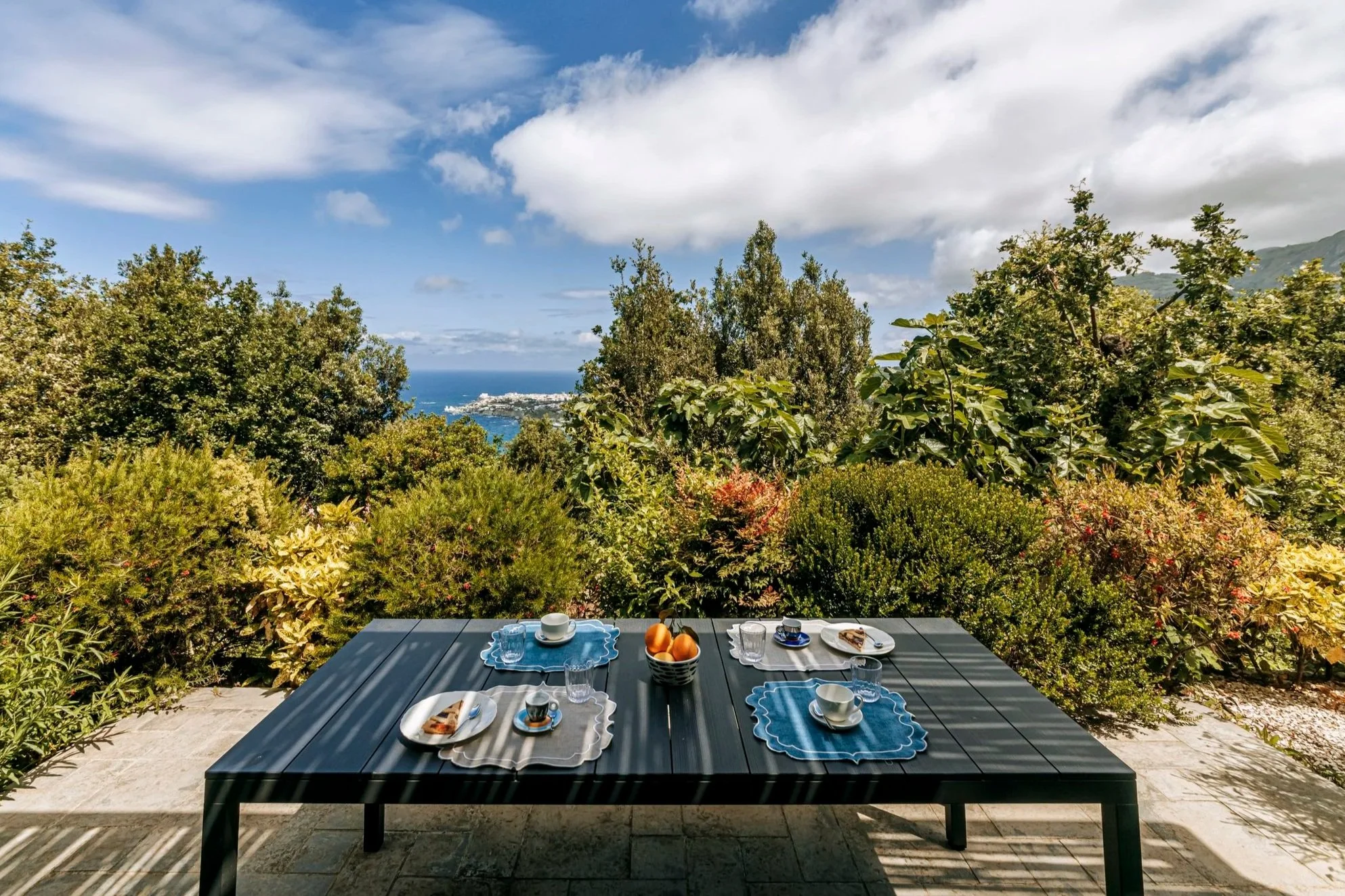 Outdoor table set for breakfast with plates, cups, glasses, a bowl of oranges, and slices of cake, overlooking lush greenery with a view of the ocean and a partly cloudy sky.