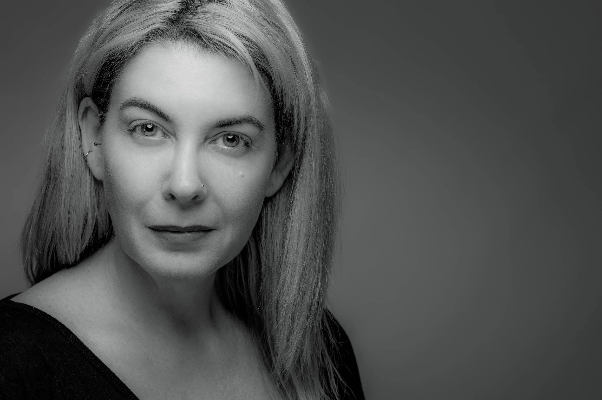 Black and white portrait of a woman with long, light-colored hair, wearing a dark top, looking directly at the camera with a neutral expression.