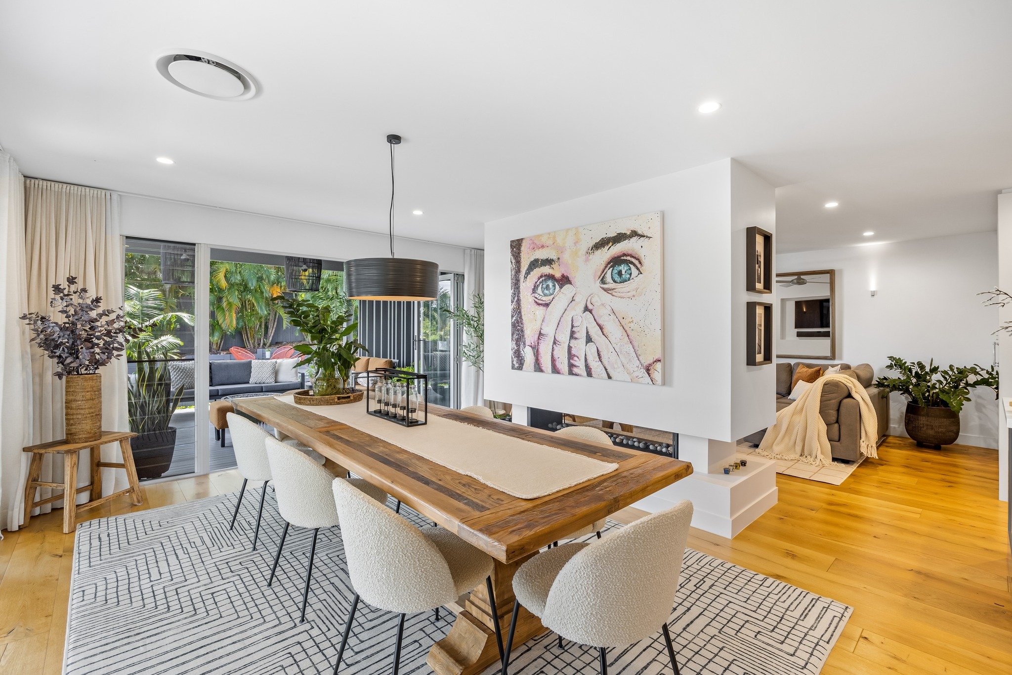 Open concept living and dining area with a wooden table, white and beige chairs, and a sliding glass door leading to a patio with outdoor seating, decorated with plants and artwork on white walls.