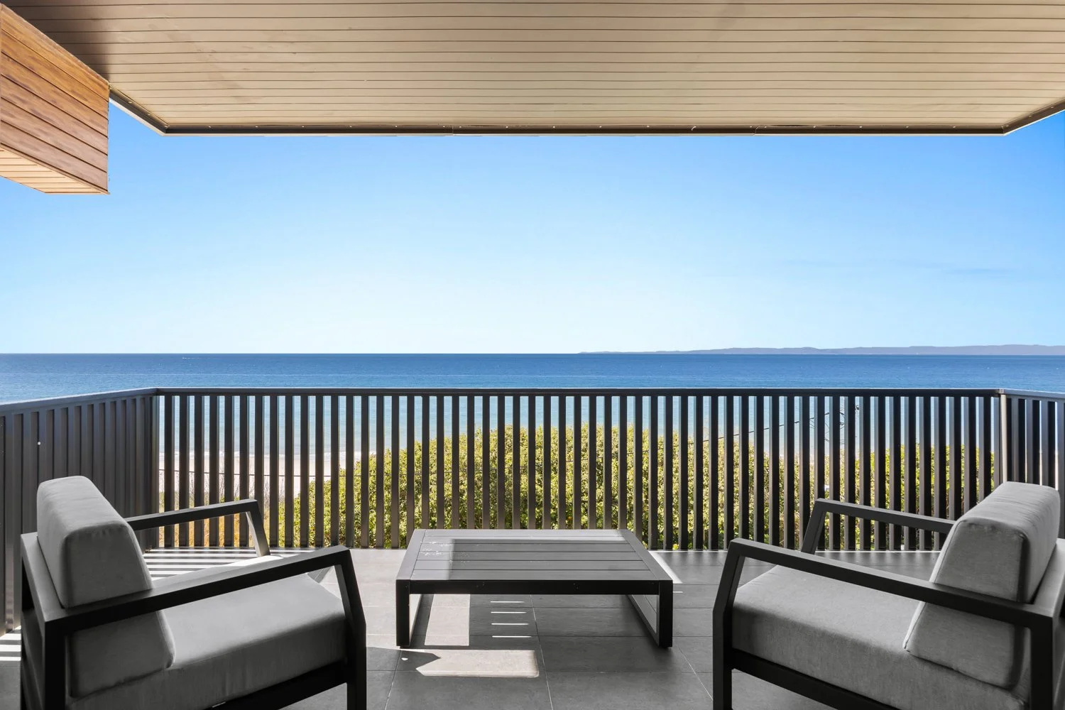 Balcony with two gray outdoor chairs and a matching coffee table, overlooking the beach and ocean under a clear blue sky.