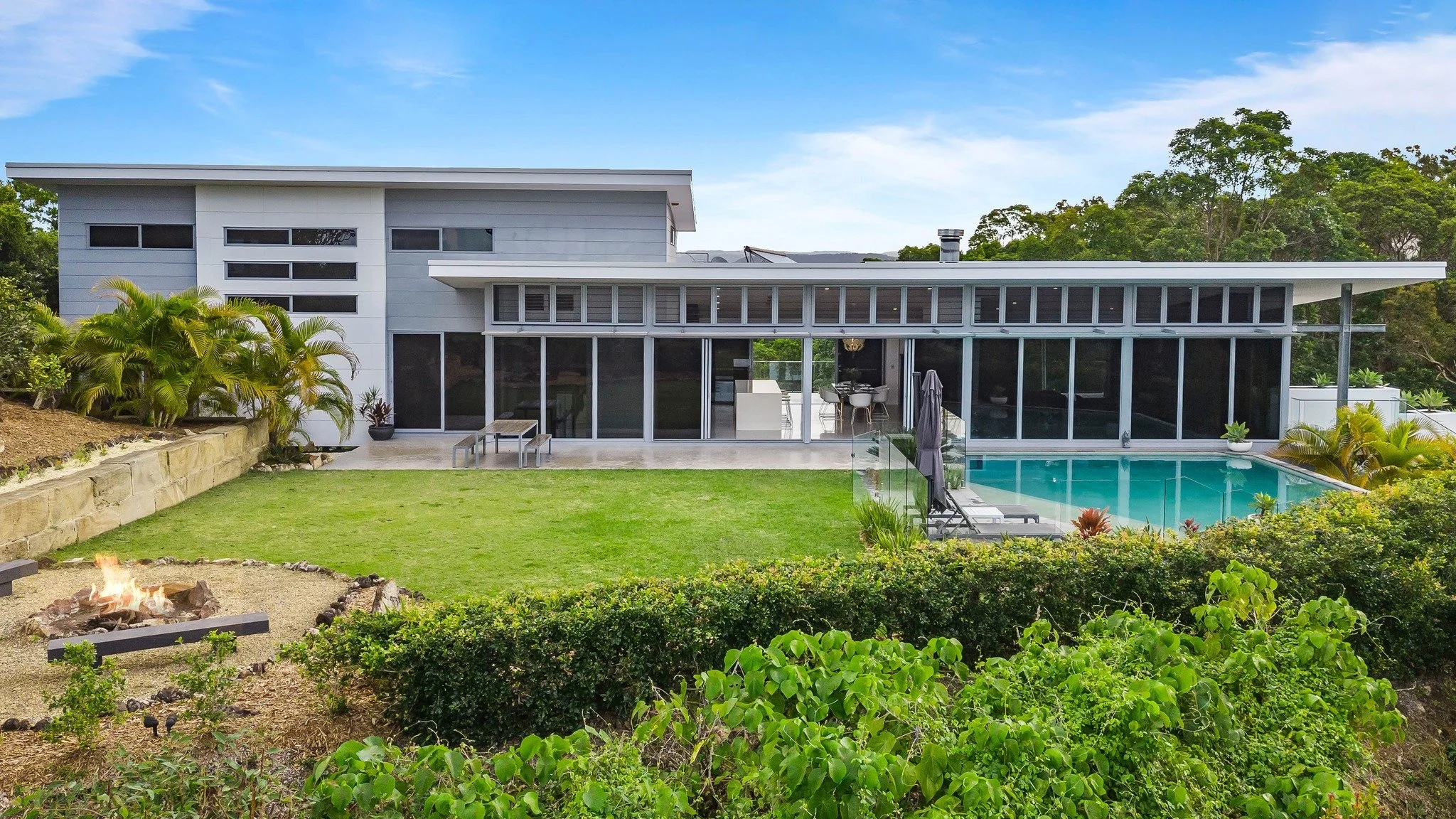 Modern house with large glass windows, outdoor pool, and lush green yard, surrounded by trees under a blue sky.