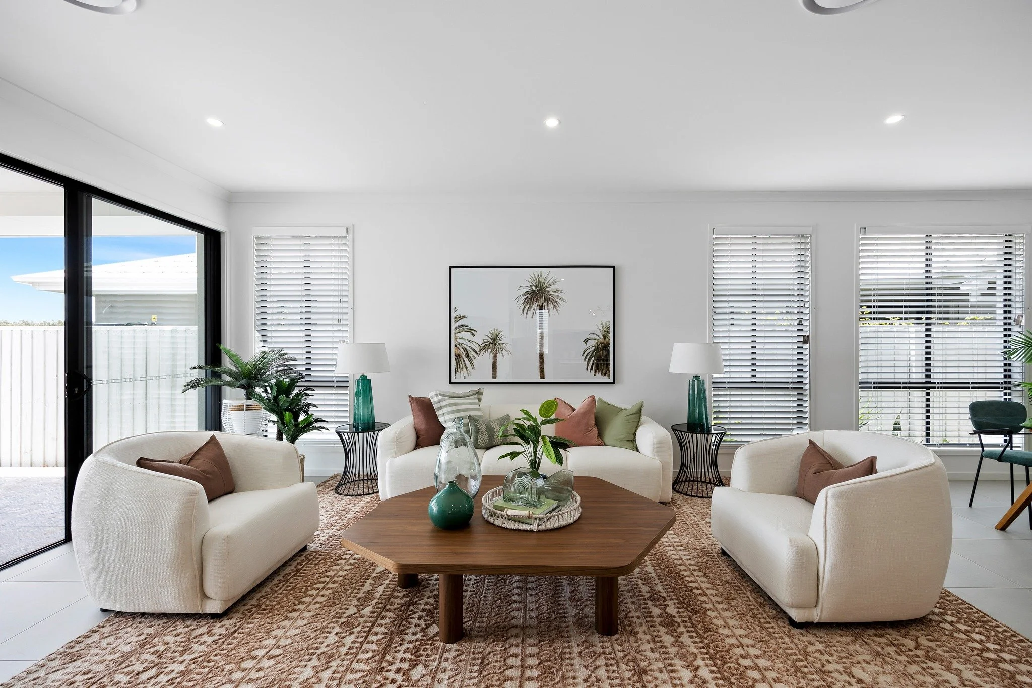 Living room with white sofas, a wooden coffee table, potted plants, and a framed palm tree print on the wall. There are three large windows with blinds and sliding glass door leading outside.