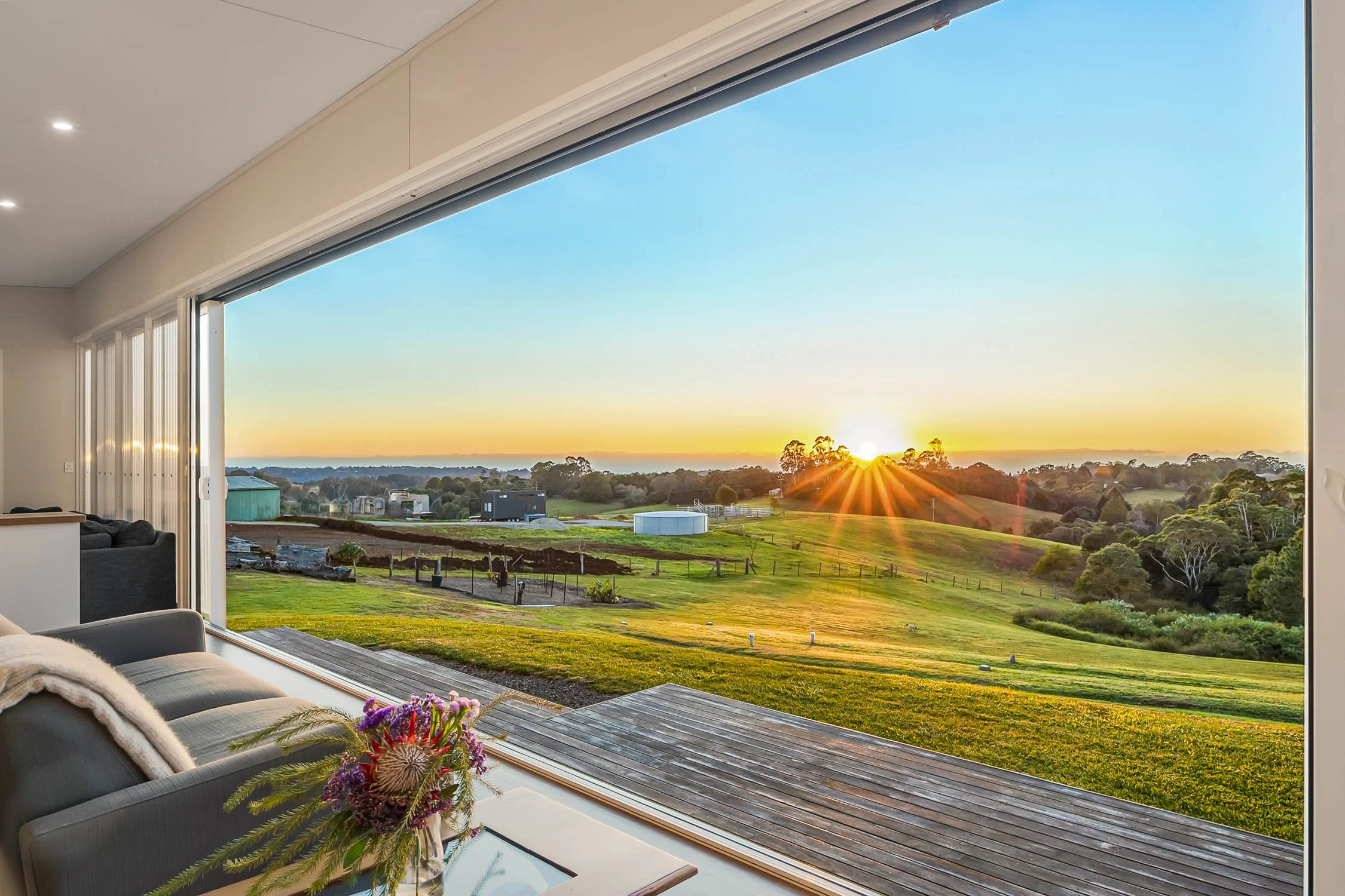 Living room with a large window overlooking a scenic landscape during sunset, with a sofa, flowers on a table, and a view of green fields and trees.
