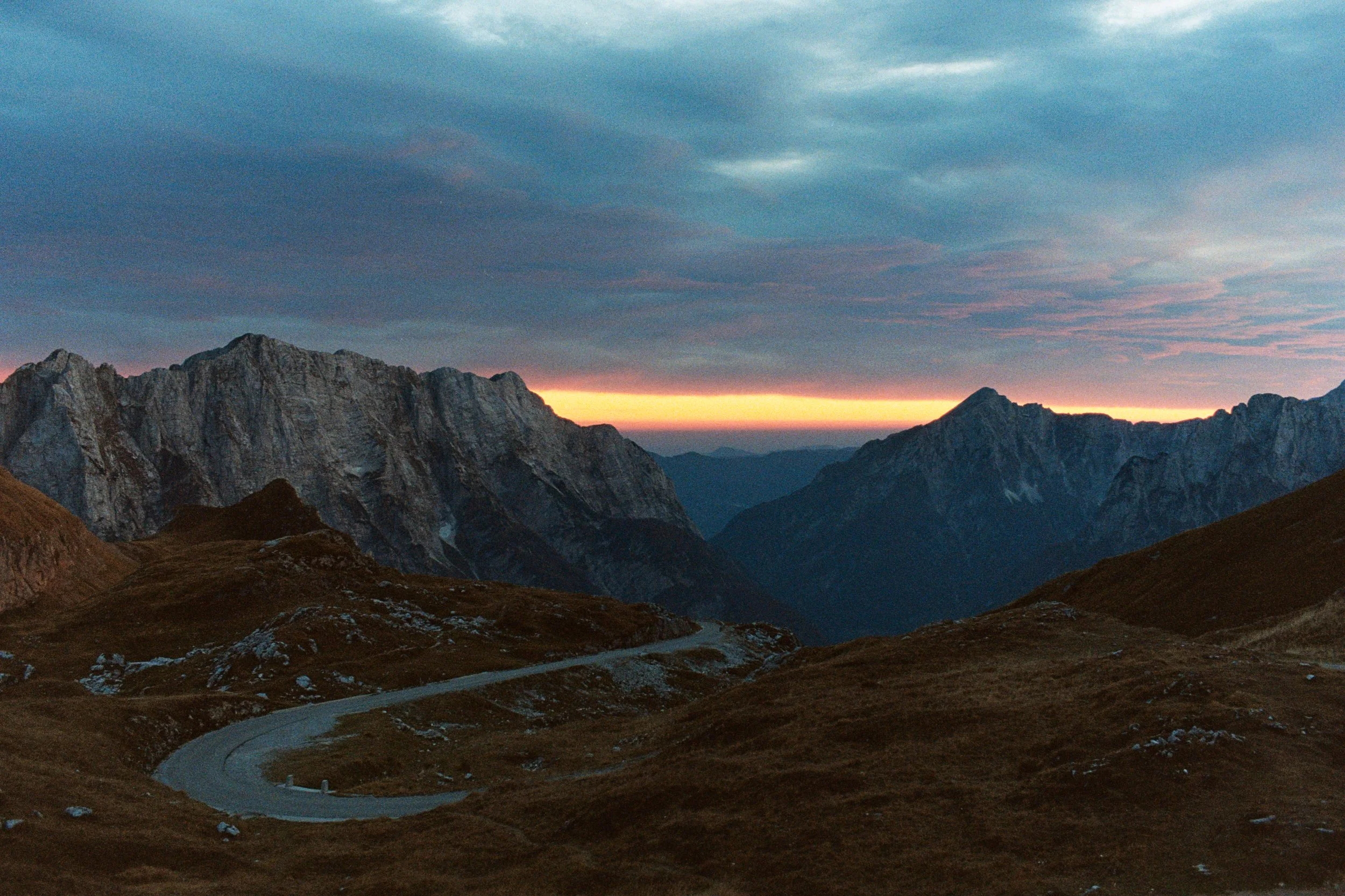 A mountain landscape at dusk with rugged peaks, a winding road in the foreground, and a colorful sky with orange and purple hues.
