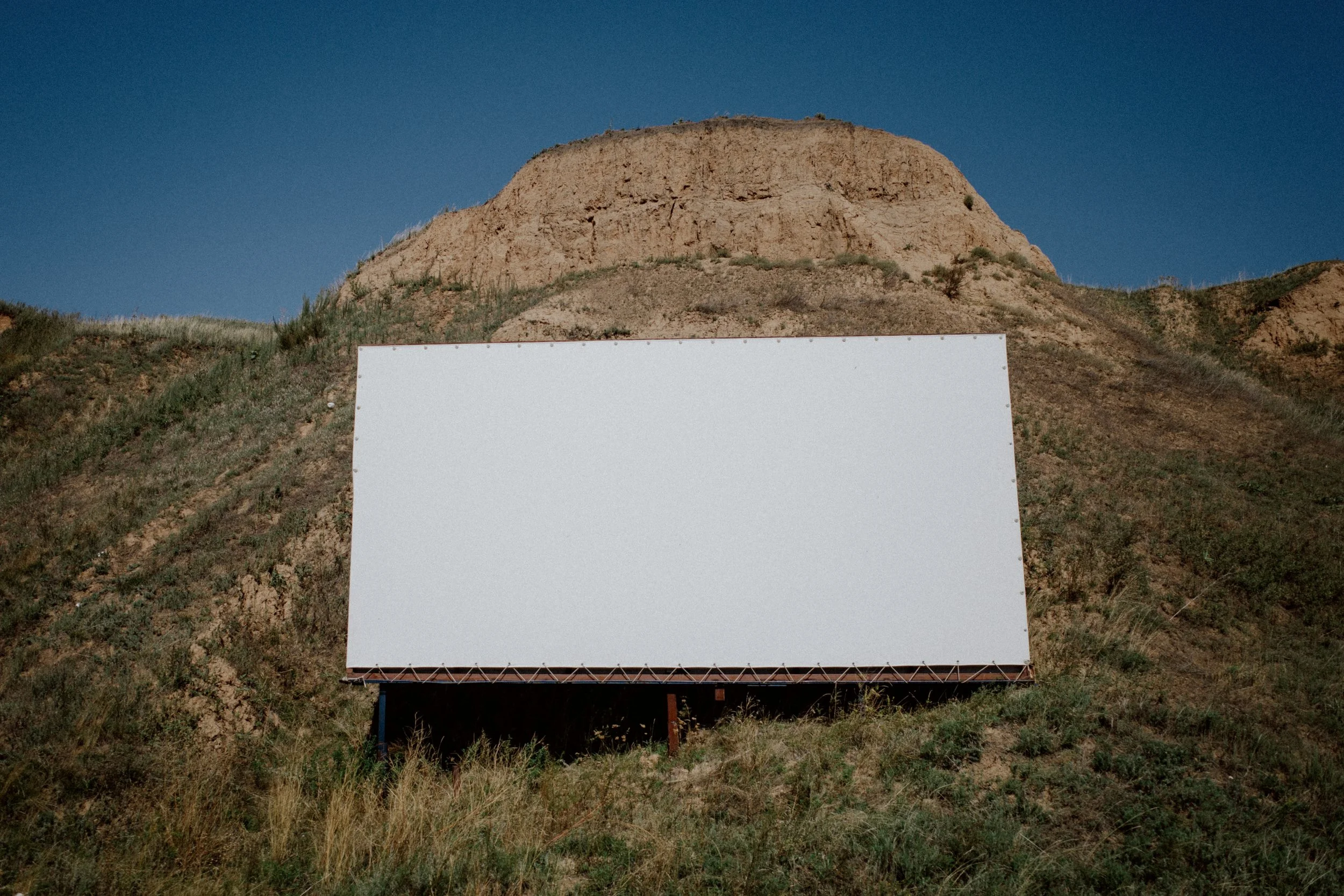 Blank white billboard on grass hill with a sandstone formation in the background under a blue sky.