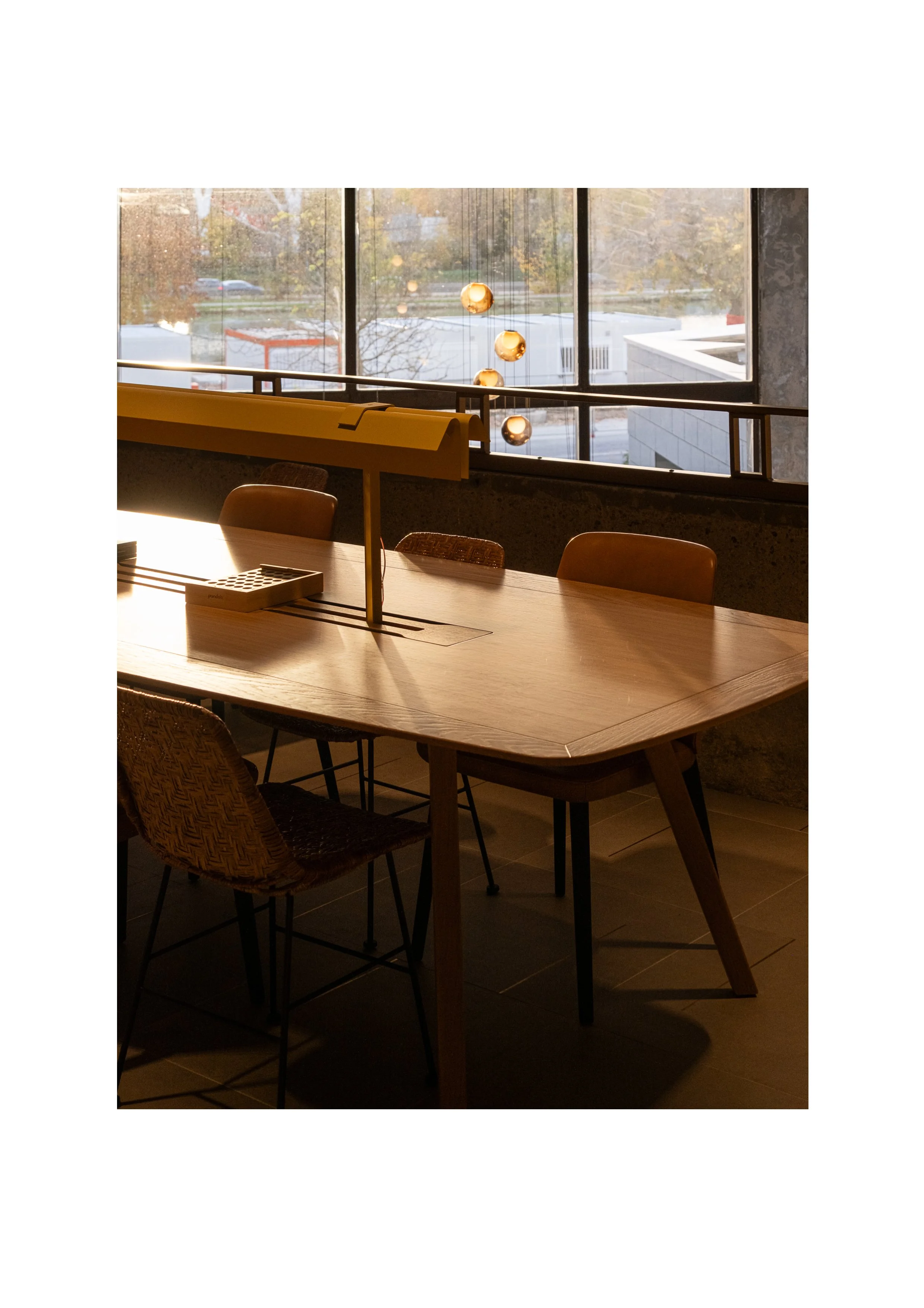 Large wooden communal table by floor-to-ceiling windows, golden hour light in a modern hotel lounge.