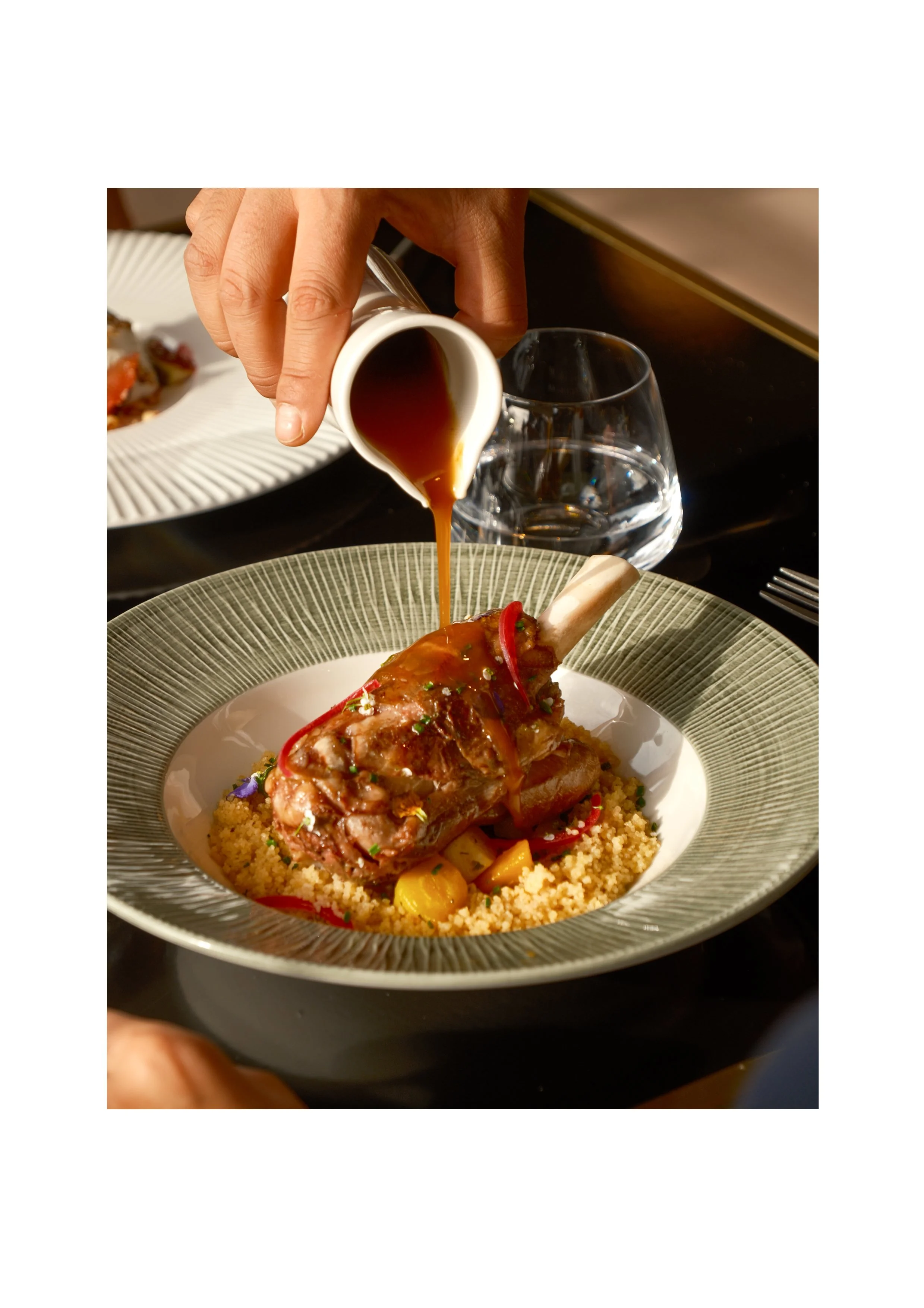 Close-up of a gourmet lamb shank being drizzled with jus, high-end culinary photography at Hyatt Centric Reims.