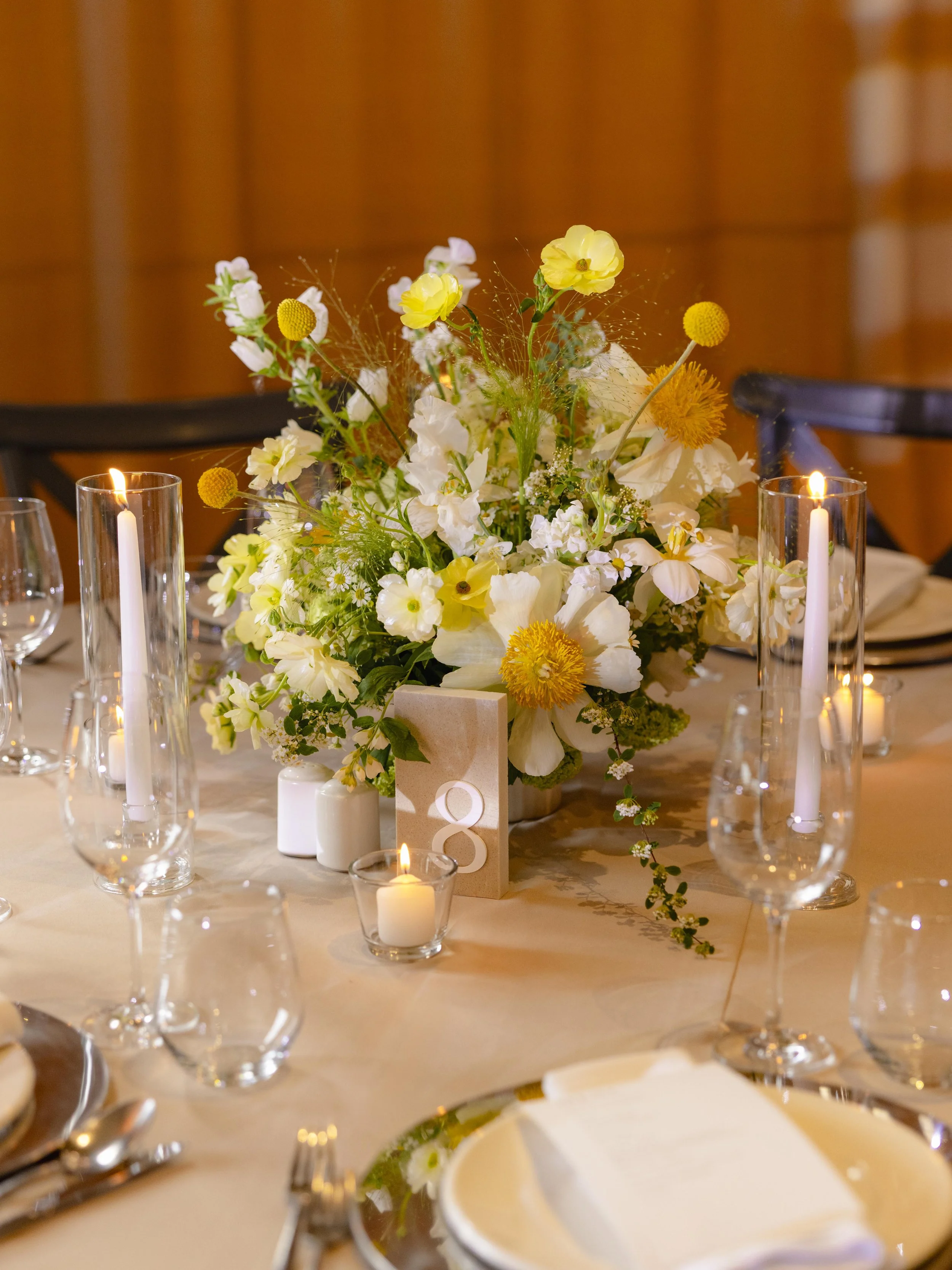 A table centerpiece with a large floral arrangement of white and yellow flowers, surrounded by tall candles in glass holders and smaller votive candles, set on an elegant table with place settings.
