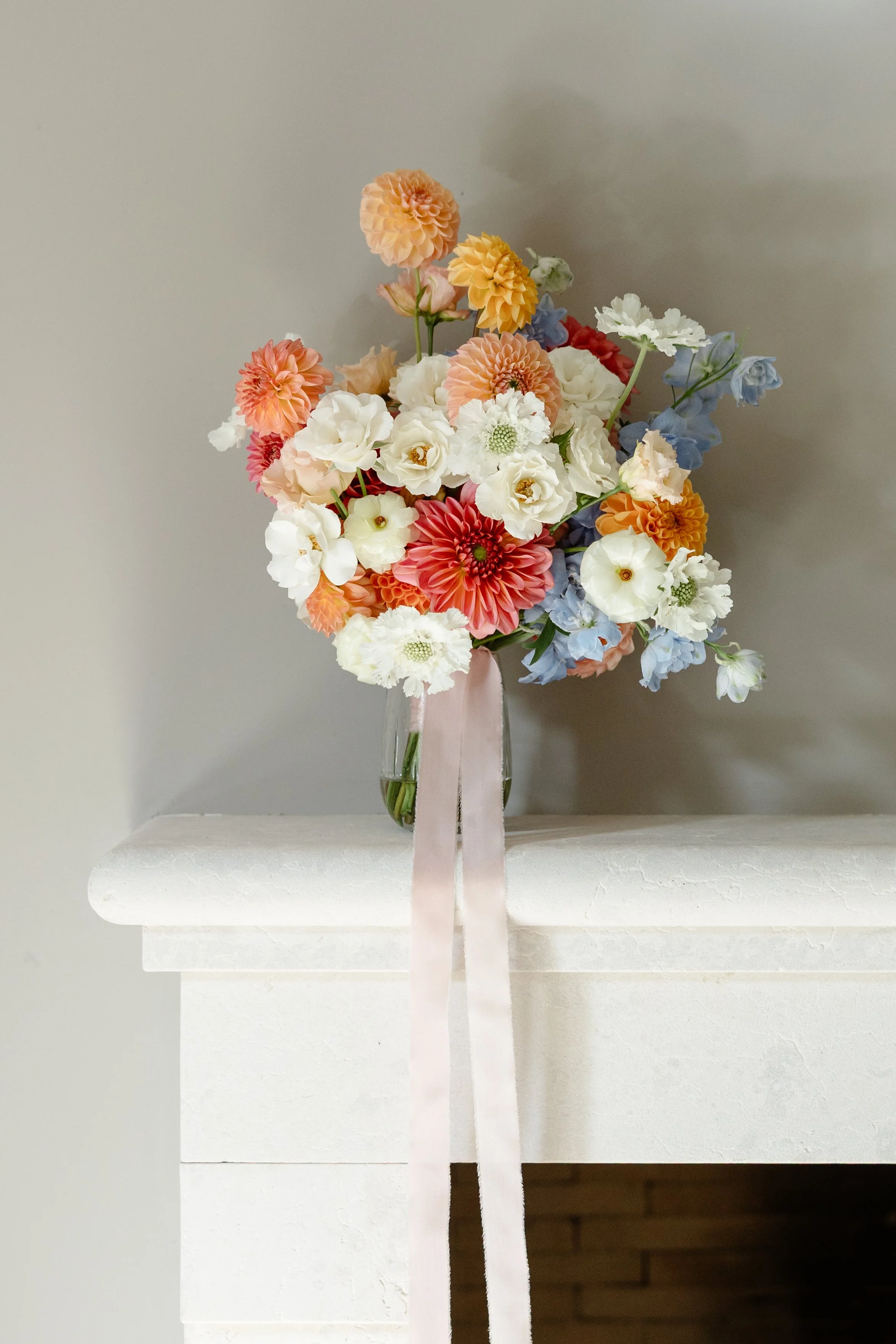 A glass vase with a pink ribbon contains a bouquet of mixed flowers, including dahlias, roses, and delphiniums, sitting on a white fireplace mantel against a plain wall.