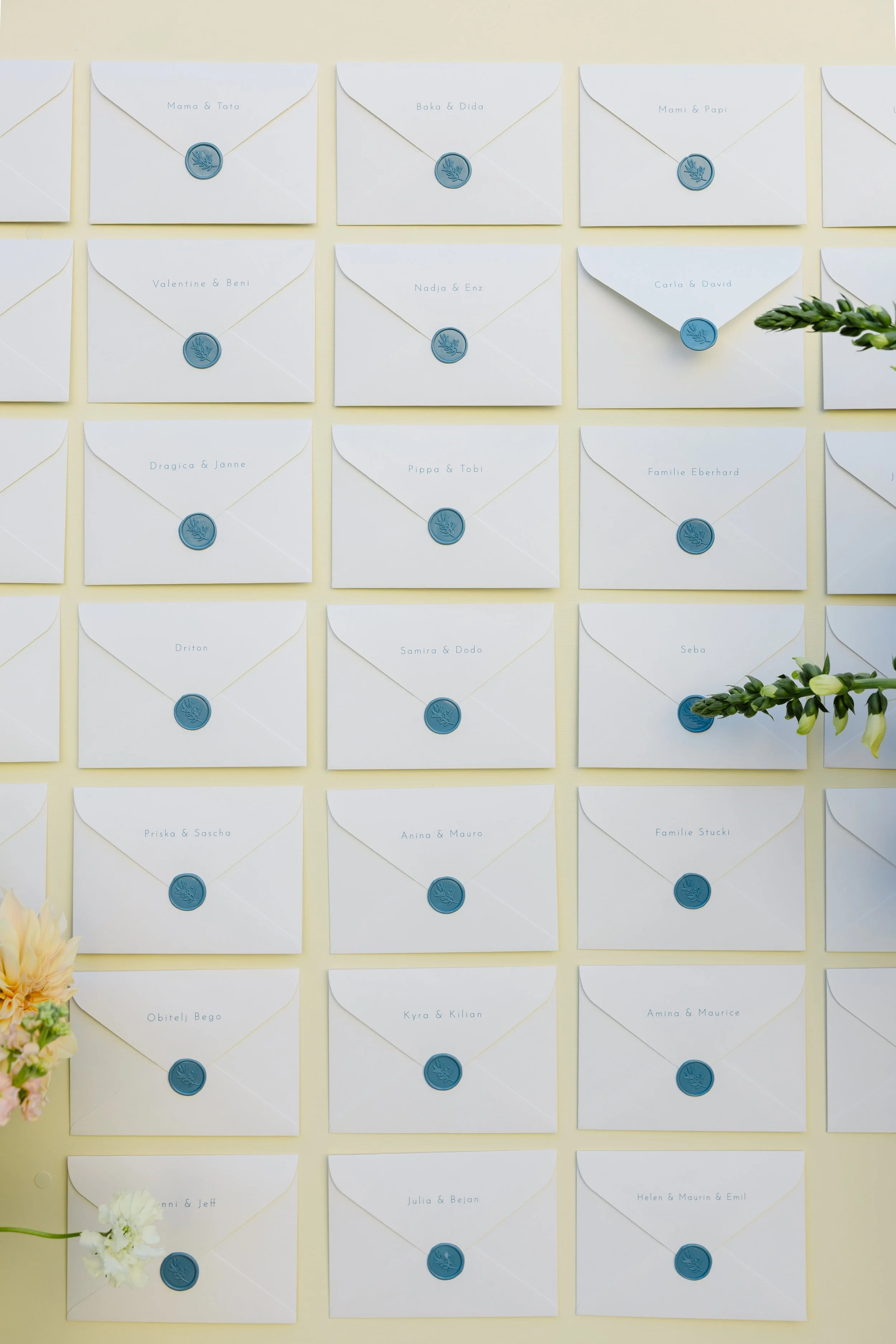Collection of white envelopes with blue wax seals, each labeled with names, arranged on a pale yellow wall, some decorated with greenery and flowers.