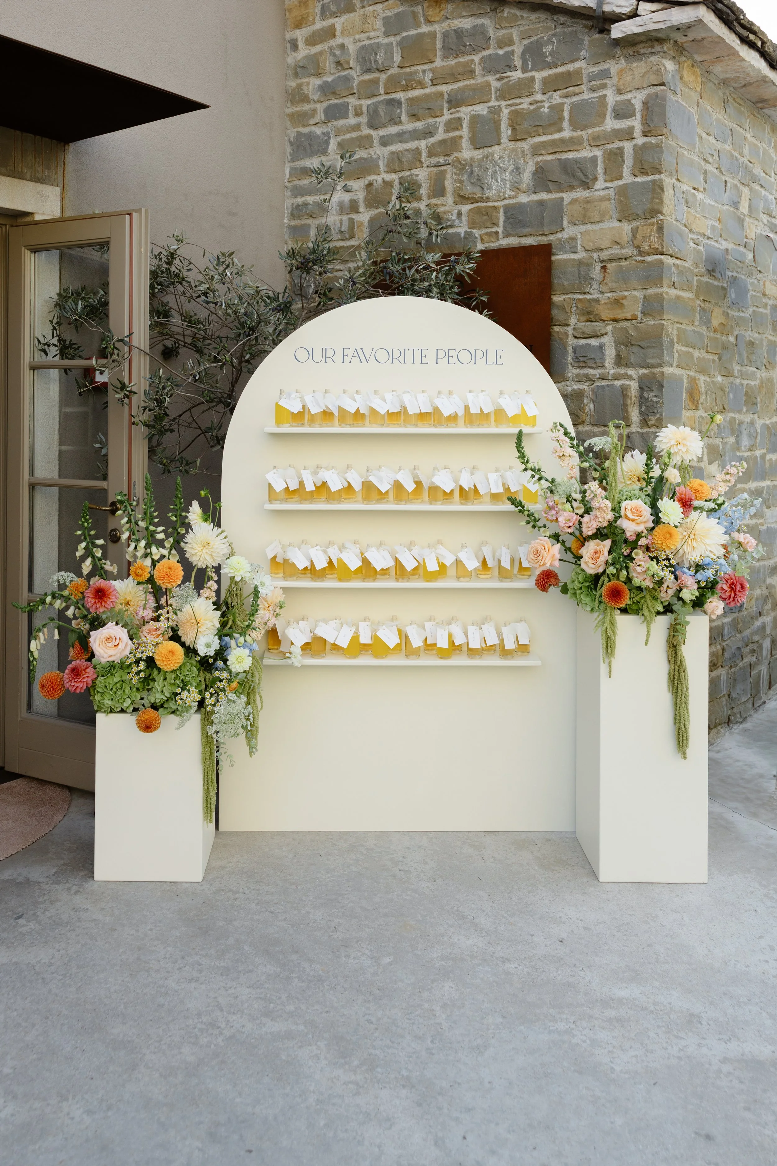 A display board with the text 'Our Favorite People' with small jars filled with honey or jam arranged on shelves beneath the text, flanked by two large floral arrangements in white planters on a concrete floor outside a brick building.