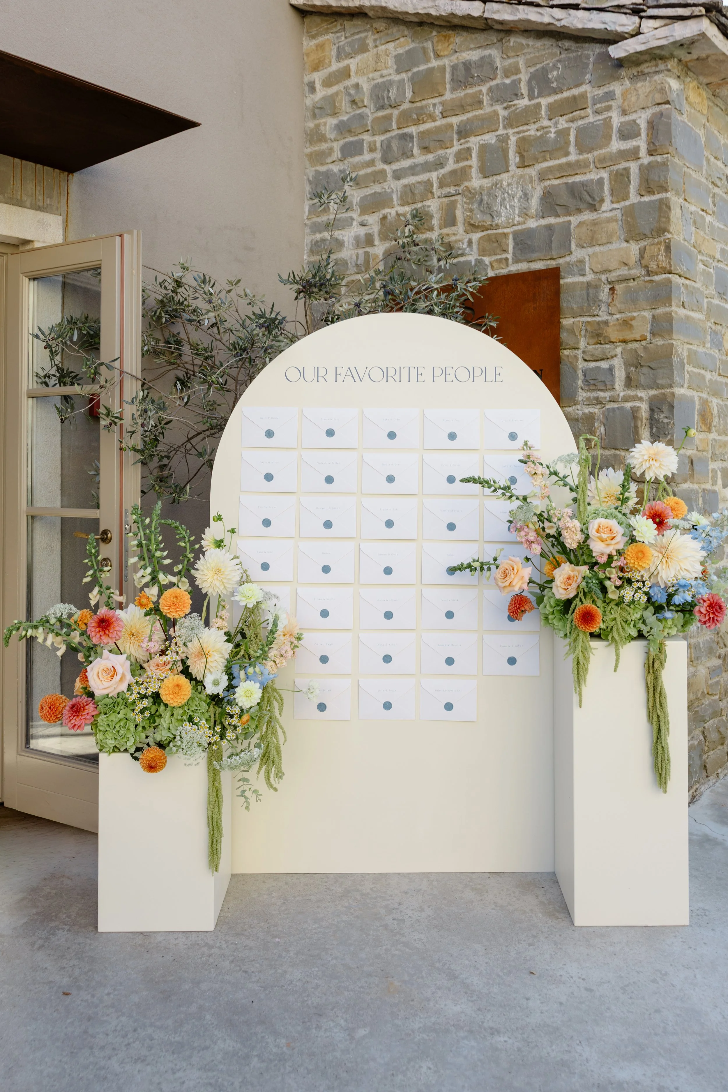 A white display board labeled 'Our Favorite People' with multiple envelopes attached, flanked by two large floral arrangements in white planters with colorful flowers, placed outside against a stone wall.