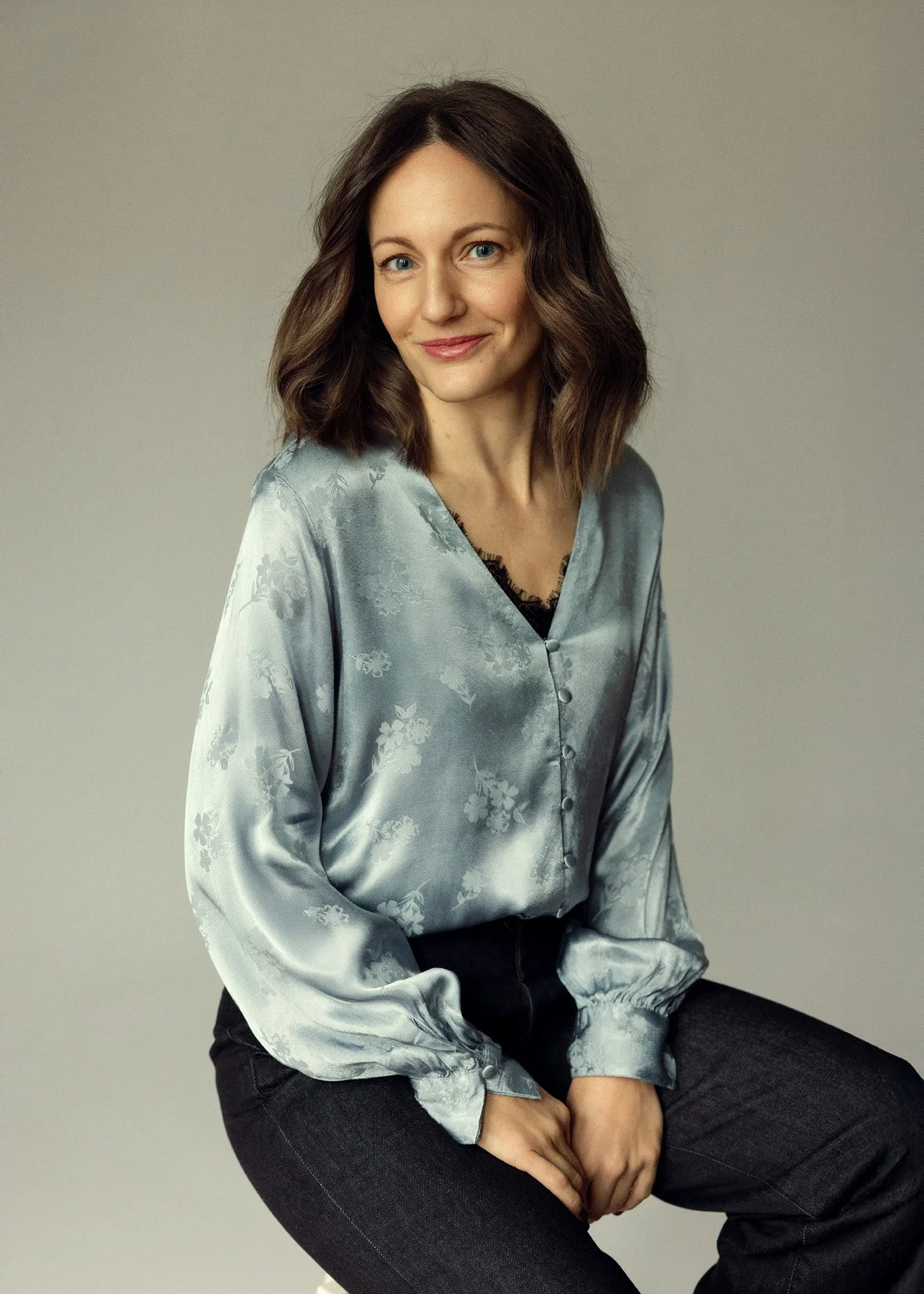 A woman with shoulder-length brown hair, blue eyes, and fair skin, wearing a light blue satin blouse with floral patterns and black pants, sitting against a plain neutral background.