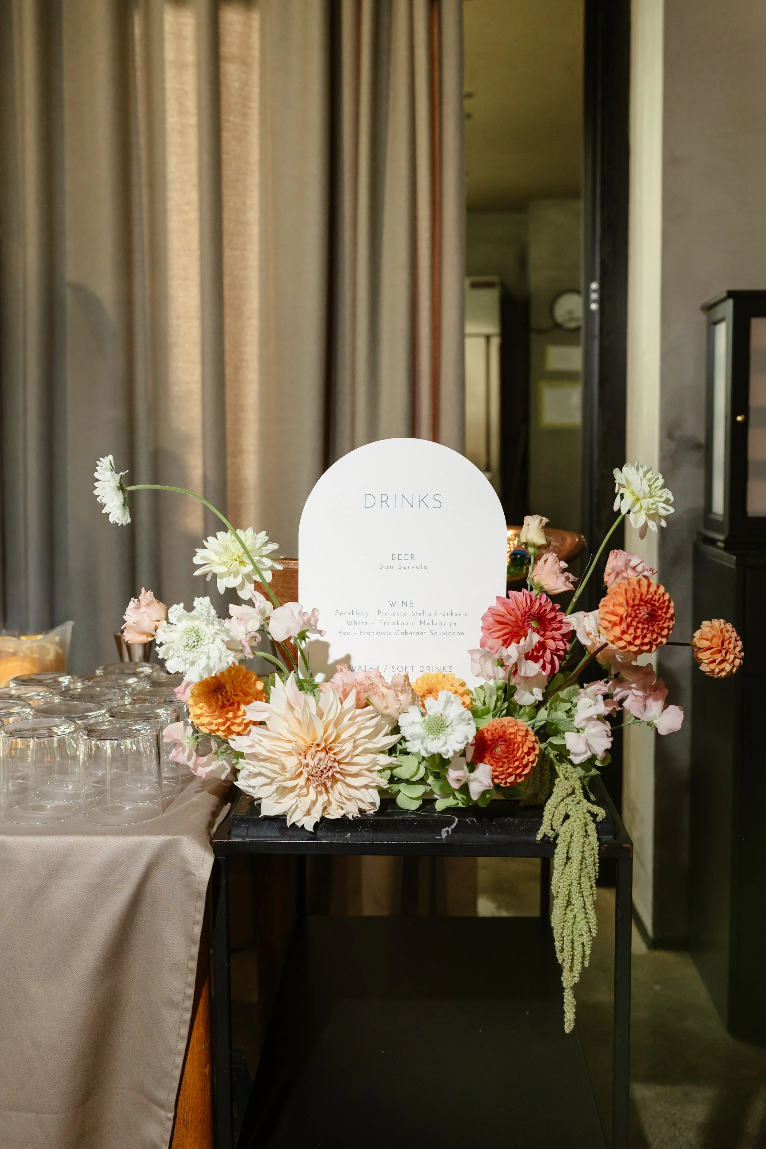 Flower arrangement with a drink menu sign on a black table near a beige tablecloth and empty glasses.