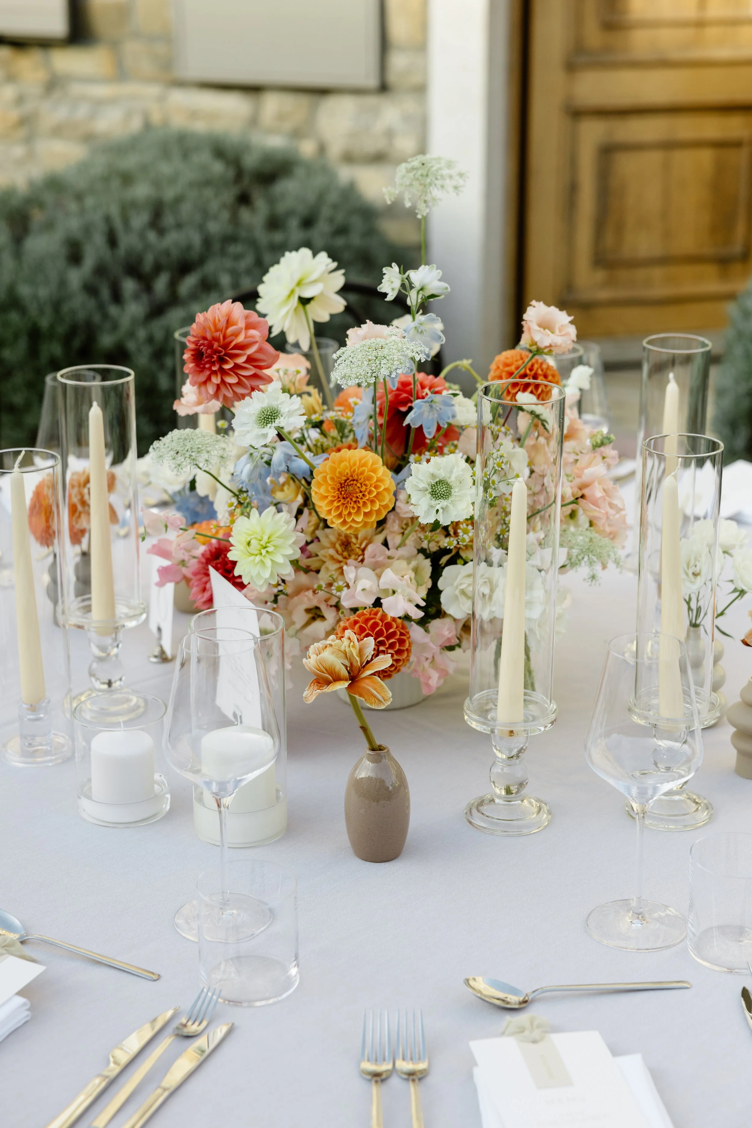 A beautifully arranged outdoor table with a white tablecloth, featuring a large colorful flower centerpiece in a vase, surrounded by tall glass candleholders with white candles, smaller candles, glassware, and gold-colored cutlery.