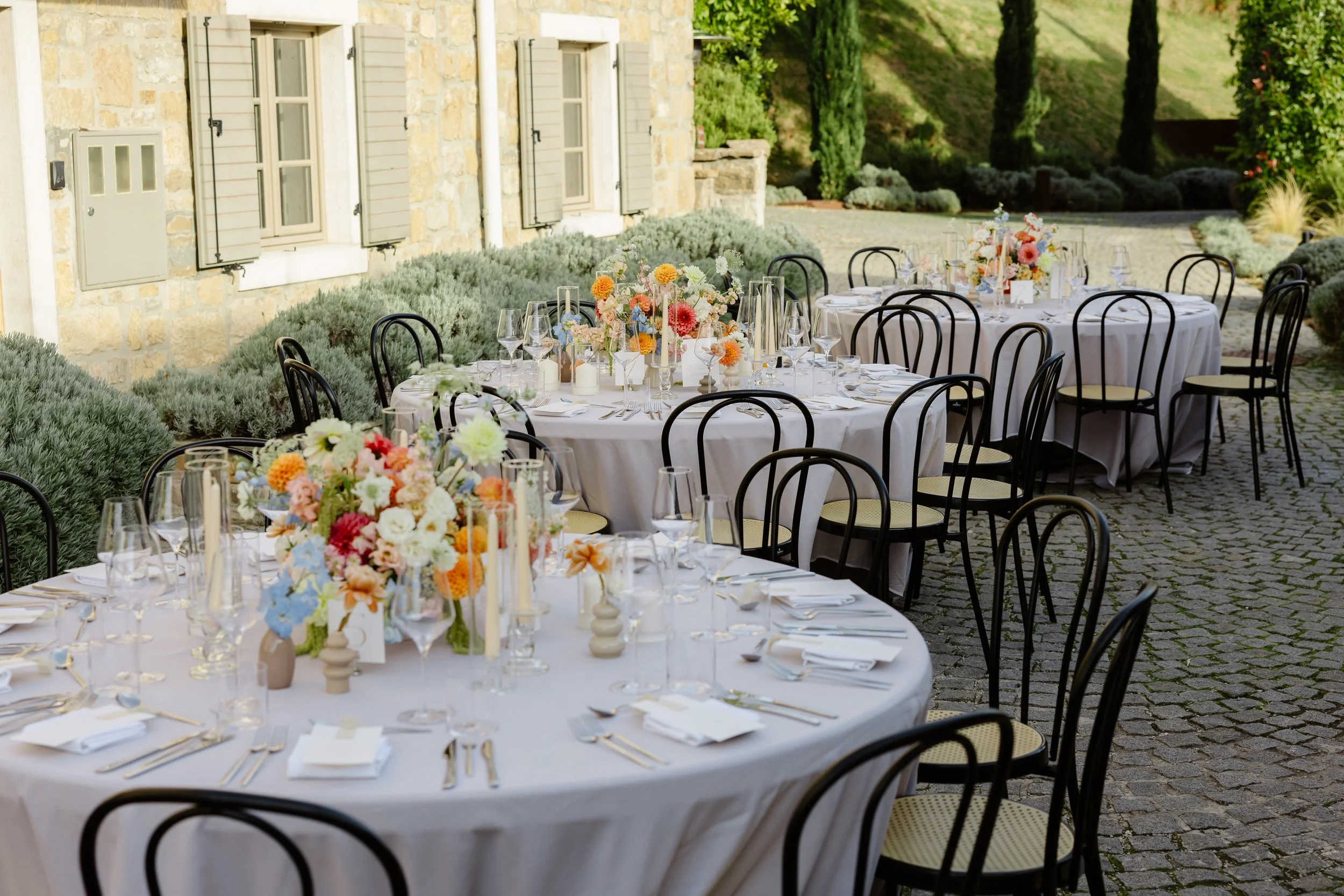 Outdoor event setup with round tables covered in white tablecloths, decorated with floral centerpieces, candles, and glassware, surrounded by black chairs on a cobblestone patio near a stone building with windows and shutters, and lush greenery in th