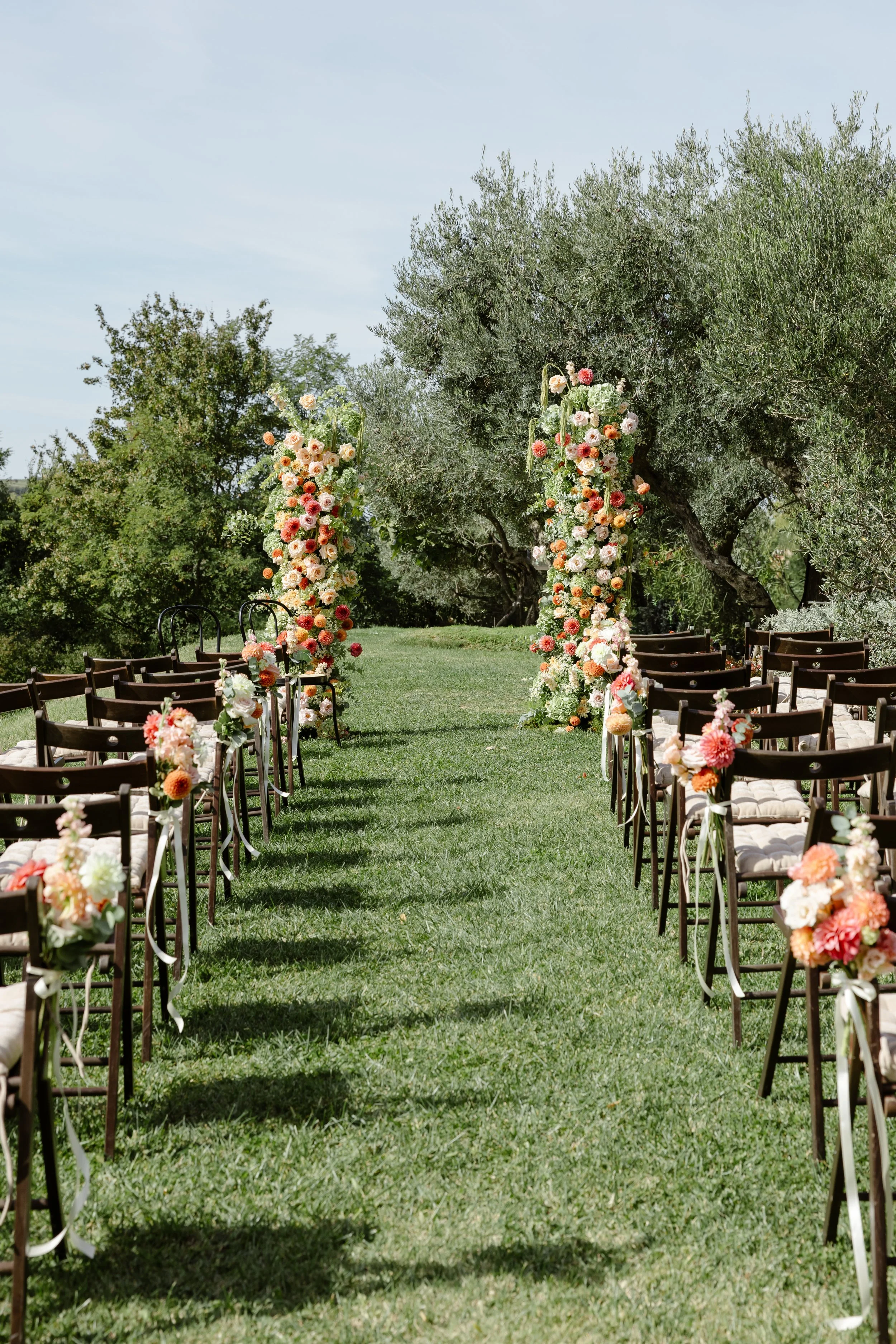 Outdoor wedding ceremony setup with rows of chairs decorated with flowers lining an aisle leading to a floral arch in a grassy area with trees in the background.