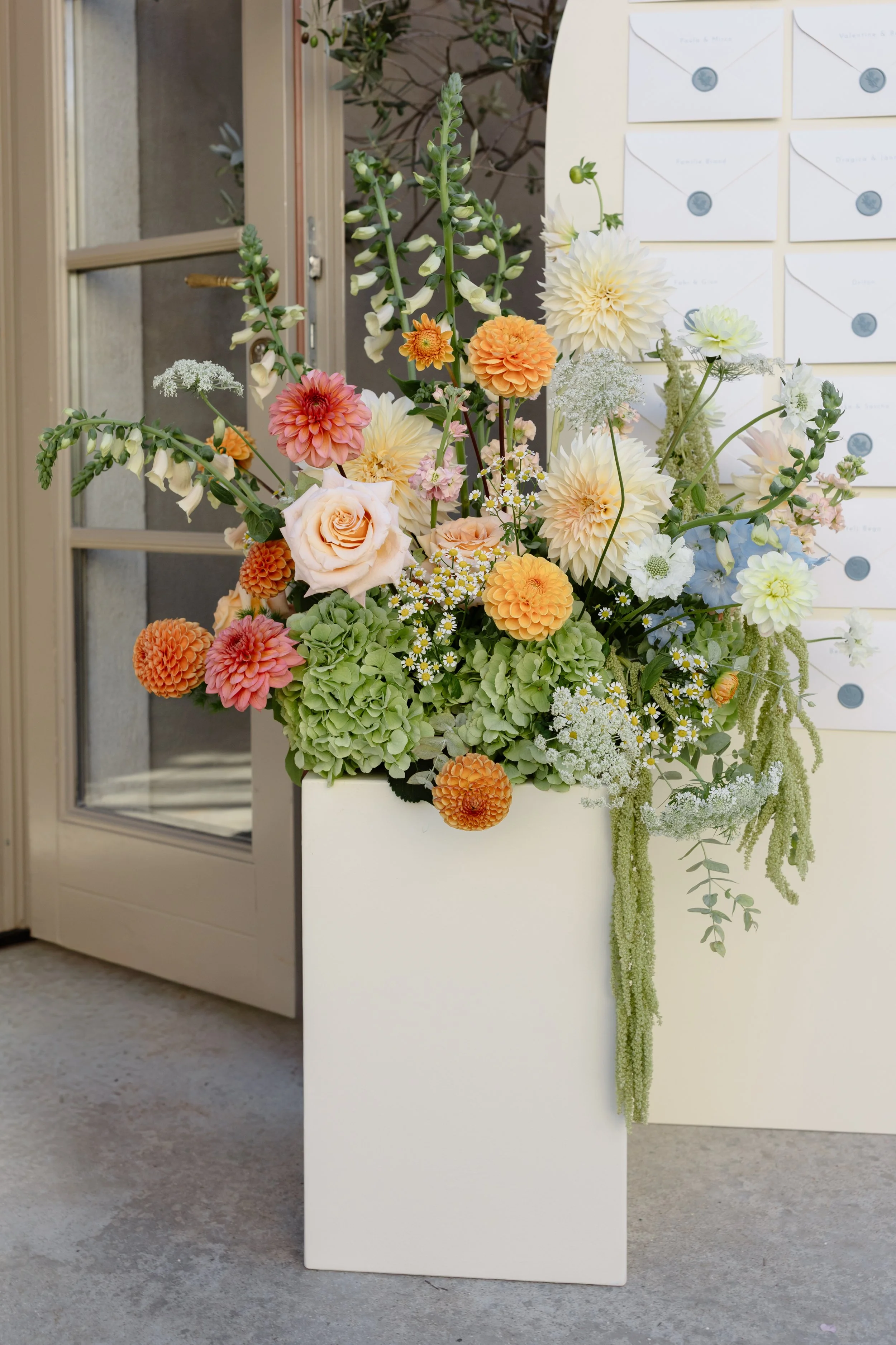 A large arrangement of various colorful flowers in a white rectangular vase near a door, with mailboxes on the wall behind.