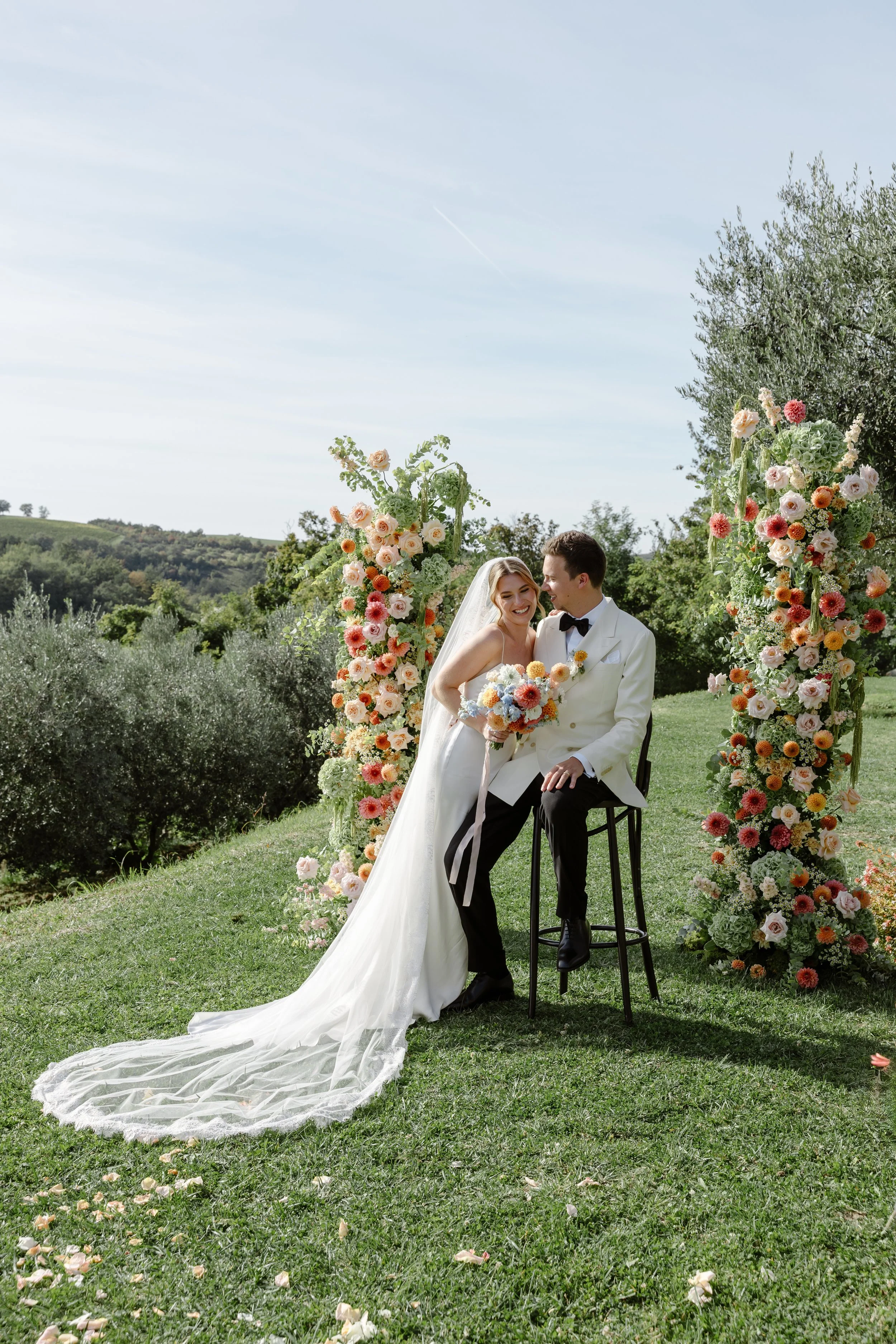 A bride and groom sitting together outdoors during their wedding ceremony, surrounded by floral decorations with a scenic green landscape in the background.