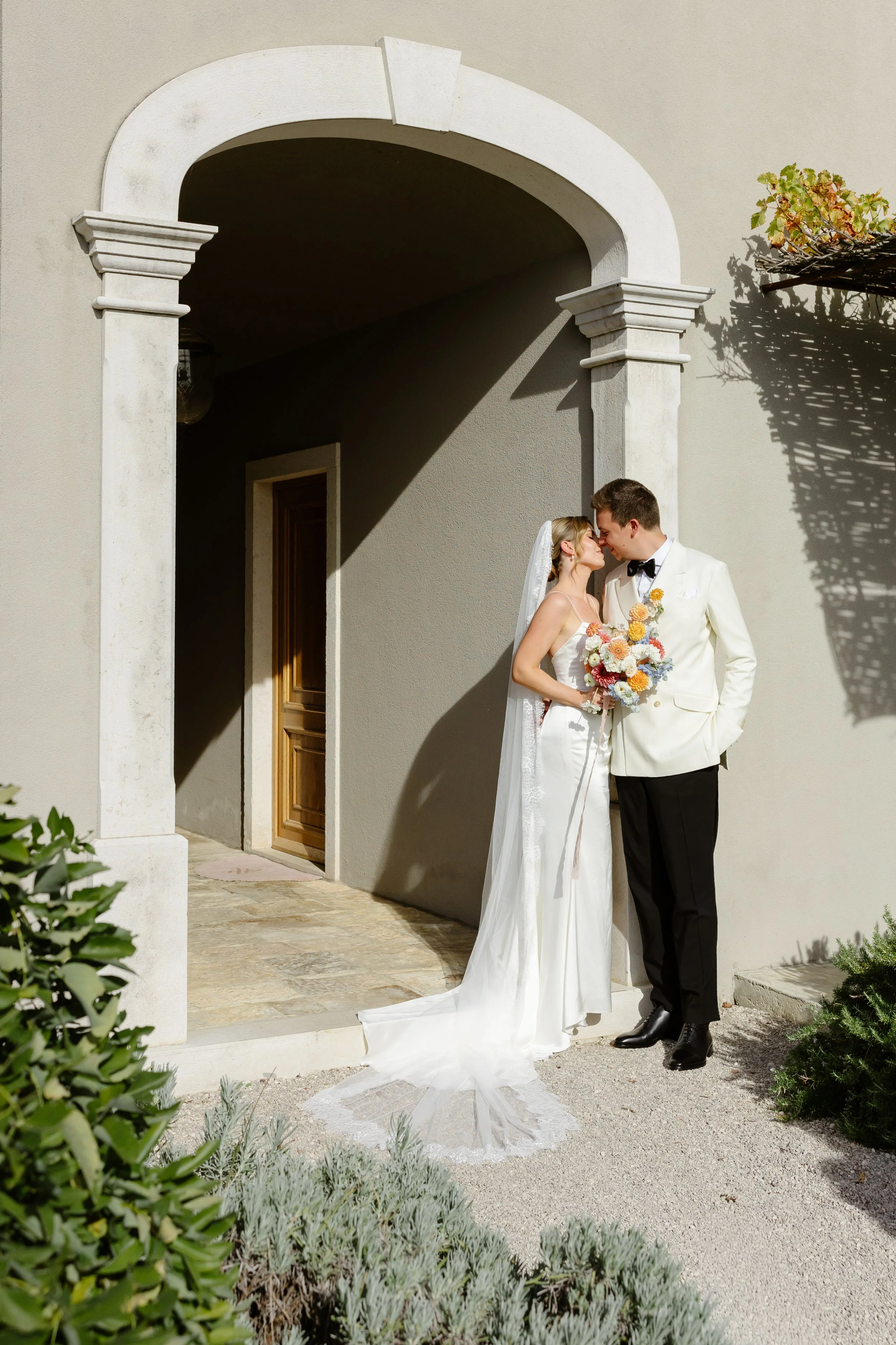 A bride and groom standing outside a building under an archway, gazing at each other, with the bride holding a colorful bouquet, in a wedding photoshoot.