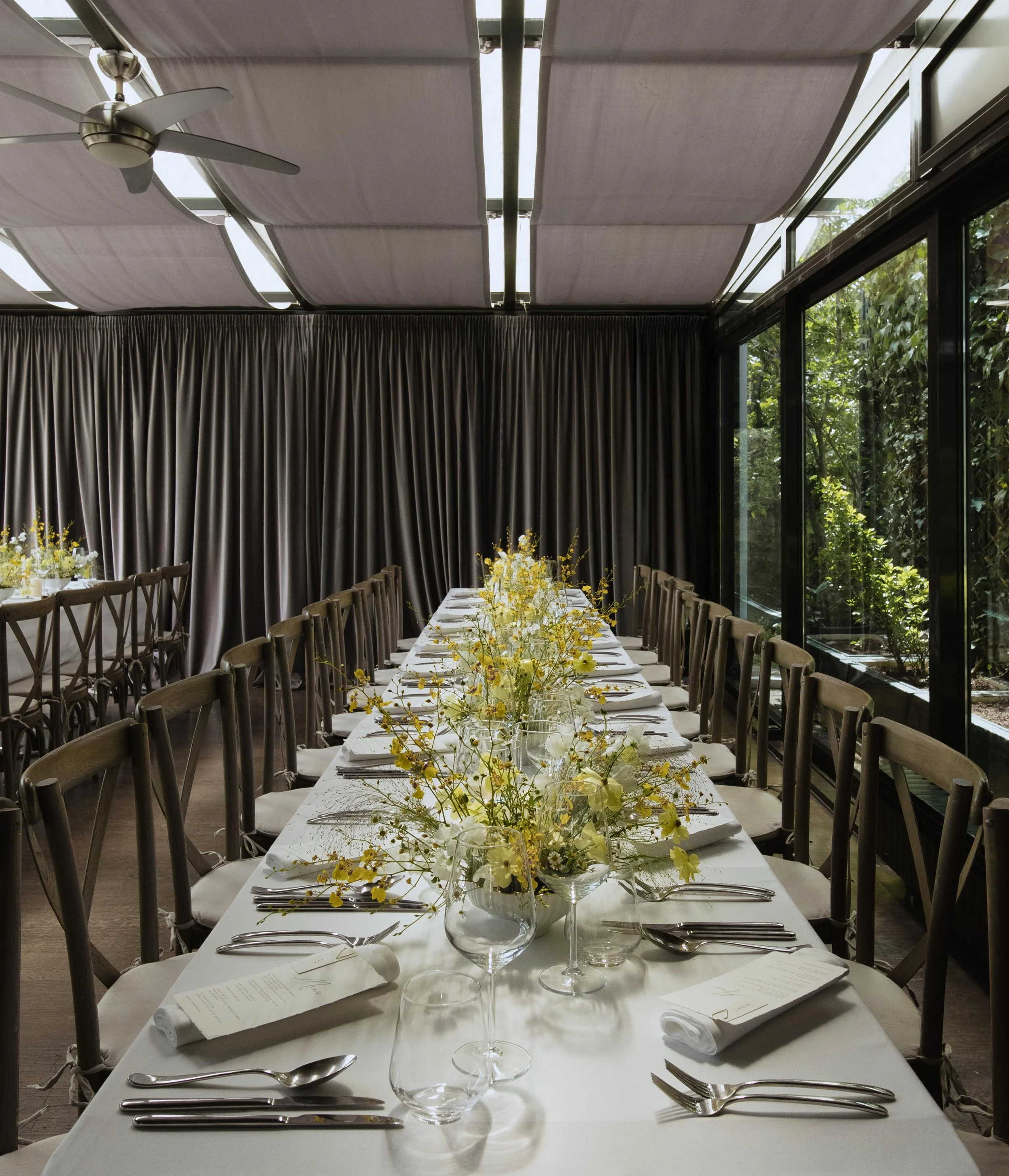 A long dining table set for a formal event with white tablecloths, floral centerpieces, wine glasses, and silverware, in a room with large windows and gray curtains.