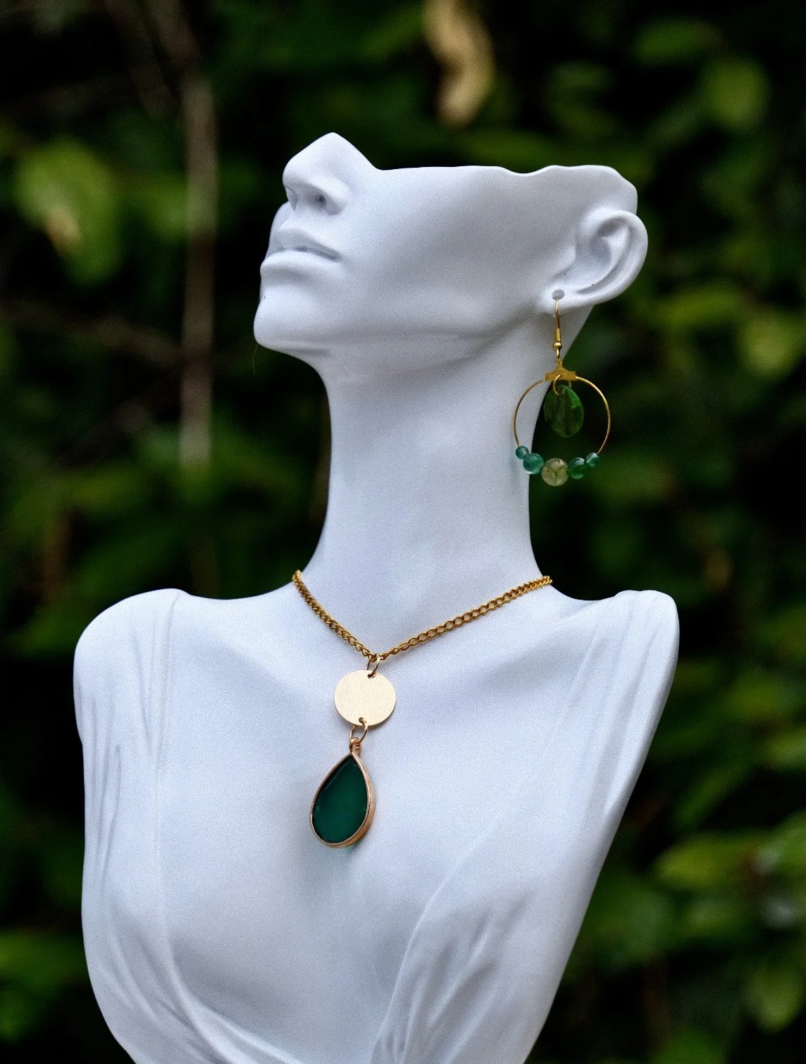 White mannequin bust displaying gold jewelry, including a necklace with a green pendant and a hoop earring with multicolored beads, set against a green foliage background.