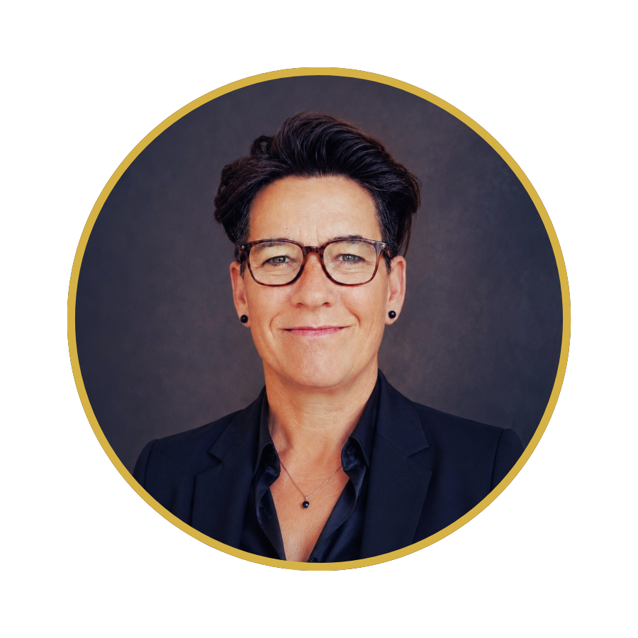 A middle-aged woman with short dark hair, glasses, and a confident smile, dressed in a black blazer and shirt, posing against a dark background.