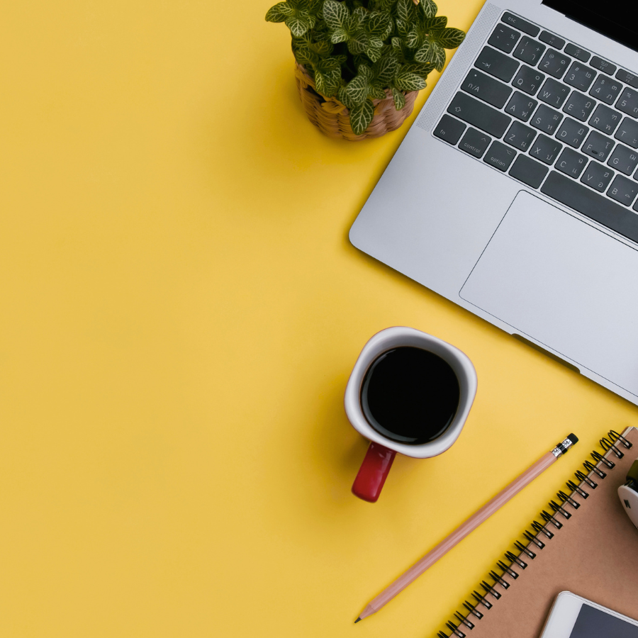 A workspace setup with a silver laptop, a small potted plant, a cup of coffee, a pink pencil, a spiral notebook with a brown cover, and a white smartphone on a yellow background.