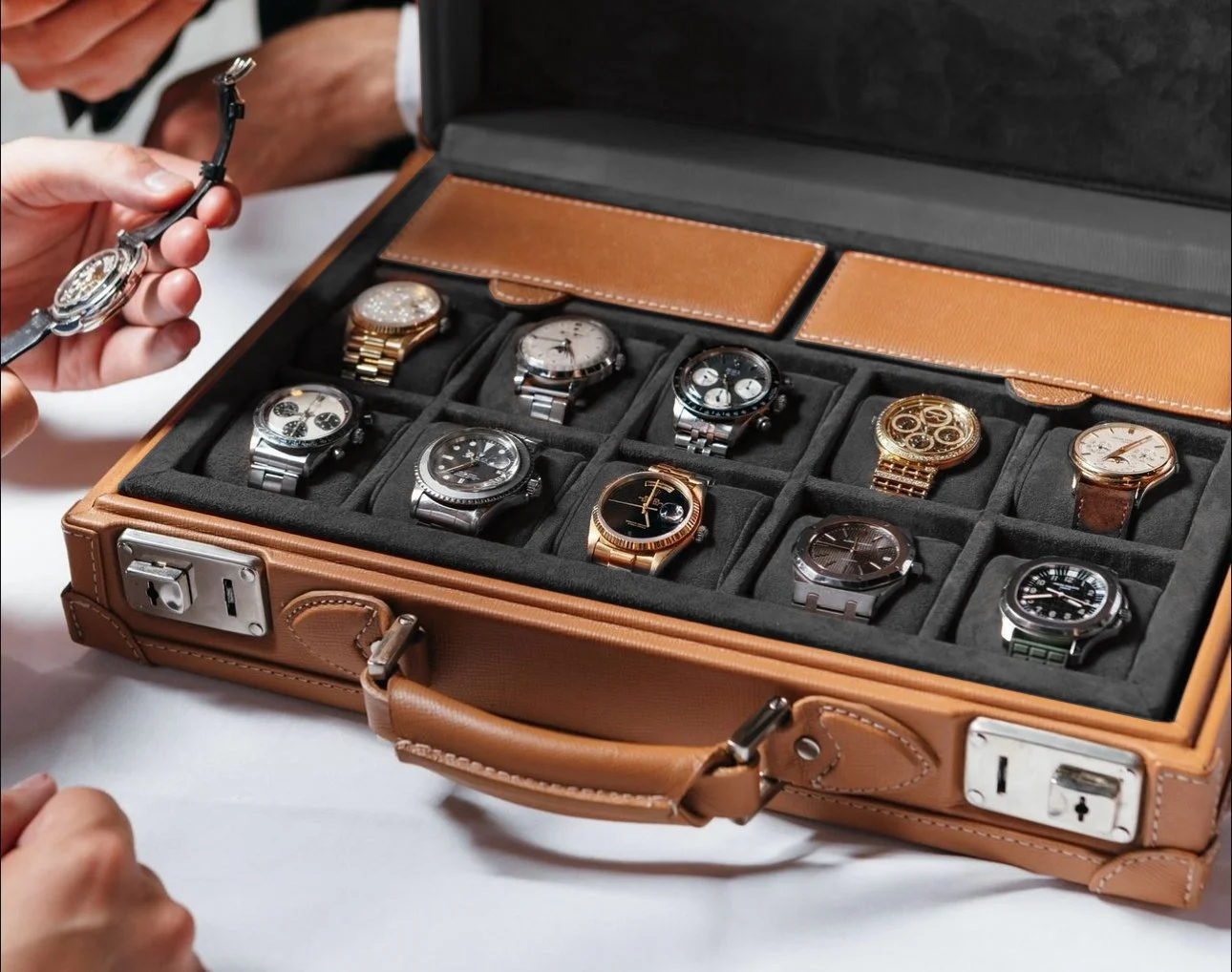 Une collection de montres de luxe dans un coffret en cuir, une personne tient une montre avec un bracelet en cuir.