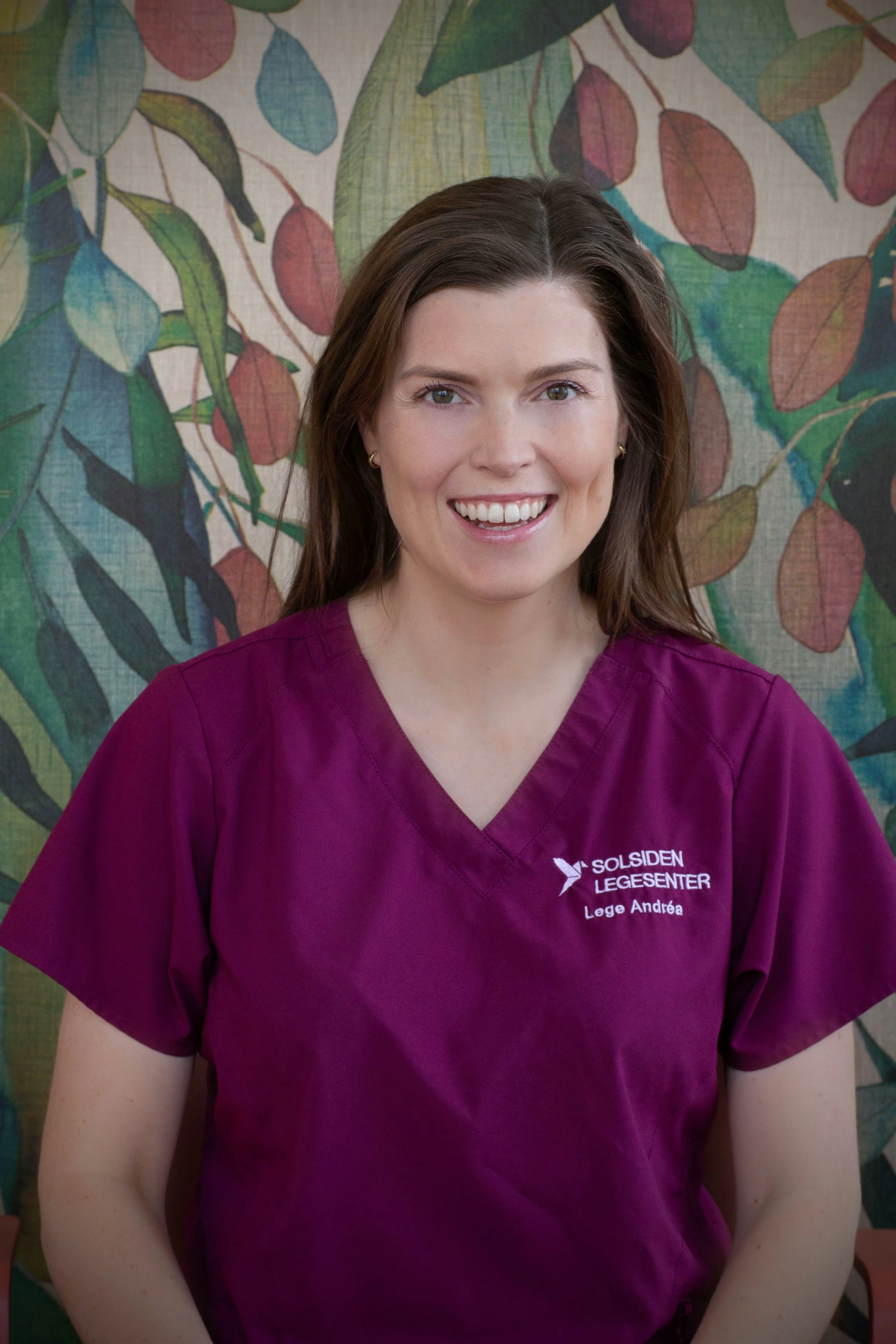 En kvinne med langt brunt hår, smilende, sitter ved et trebord i et rom med grønne planter og en glass- og trefasade i bakgrunnen.