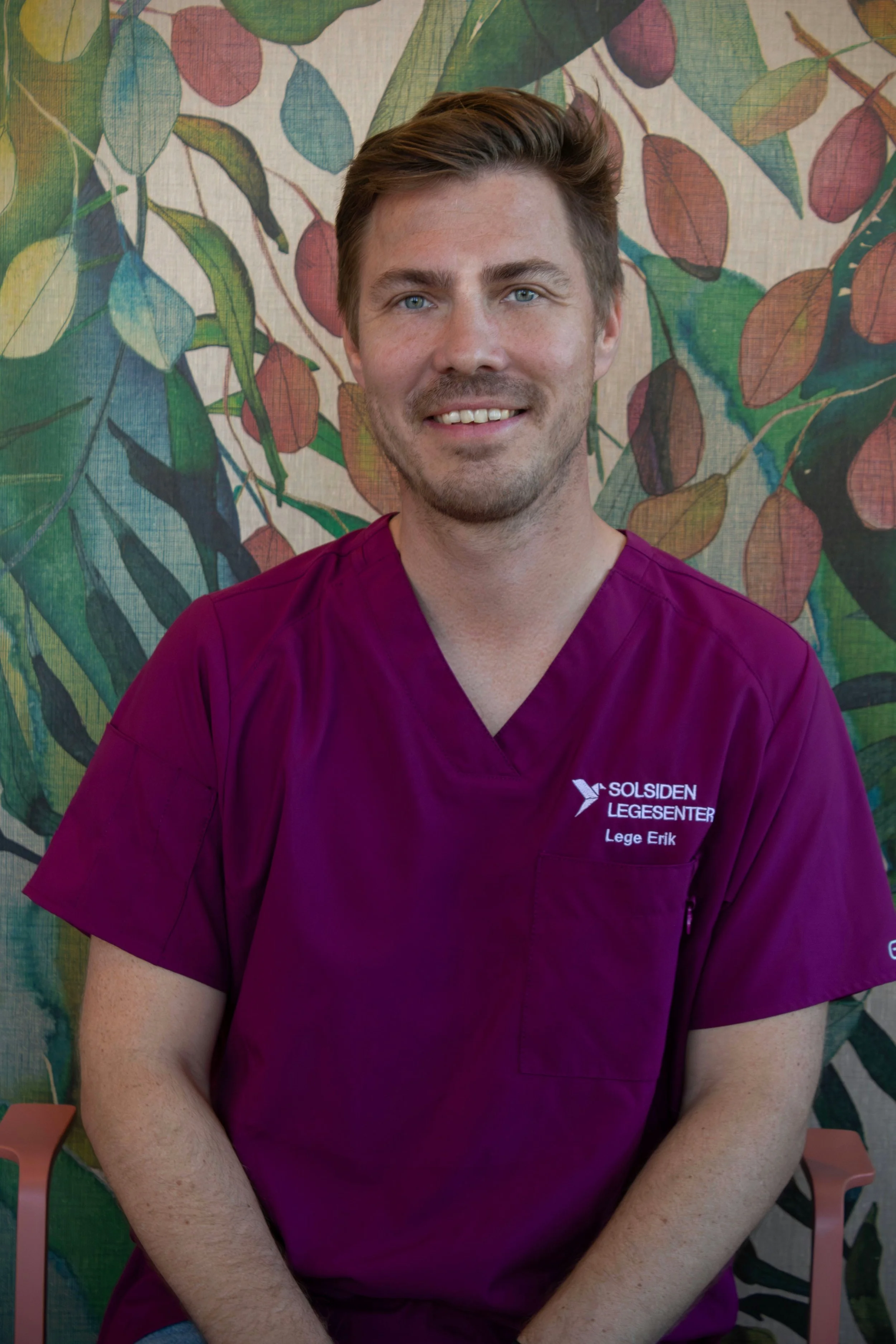 En mann med brunt hår og blått klokke smiler, sitter ved et trebord med grønne planter i bakgrunnen.