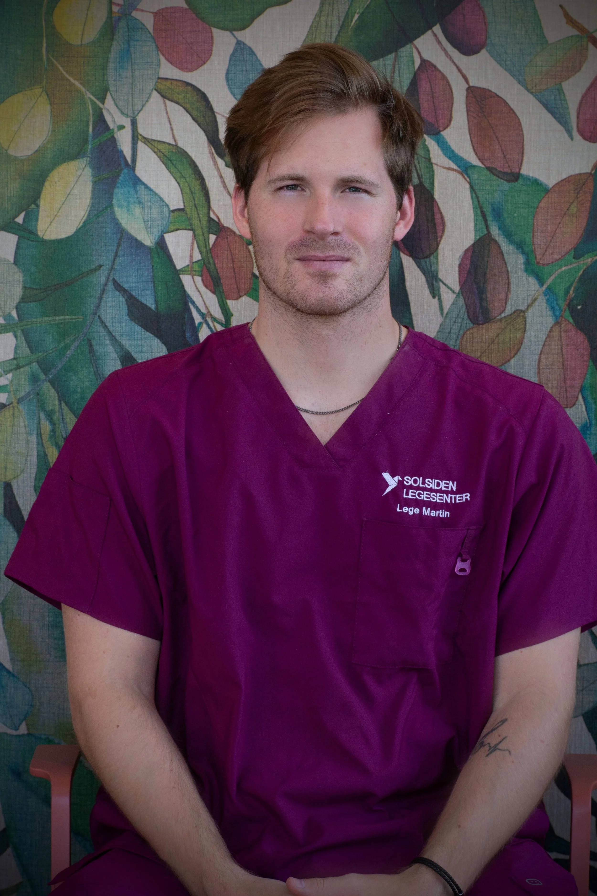 Ung mann med lyst hår og skjegg, kledd i blå genser, sitter ved et bord i et innendørs drivhus med grønne planter i bakgrunnen.