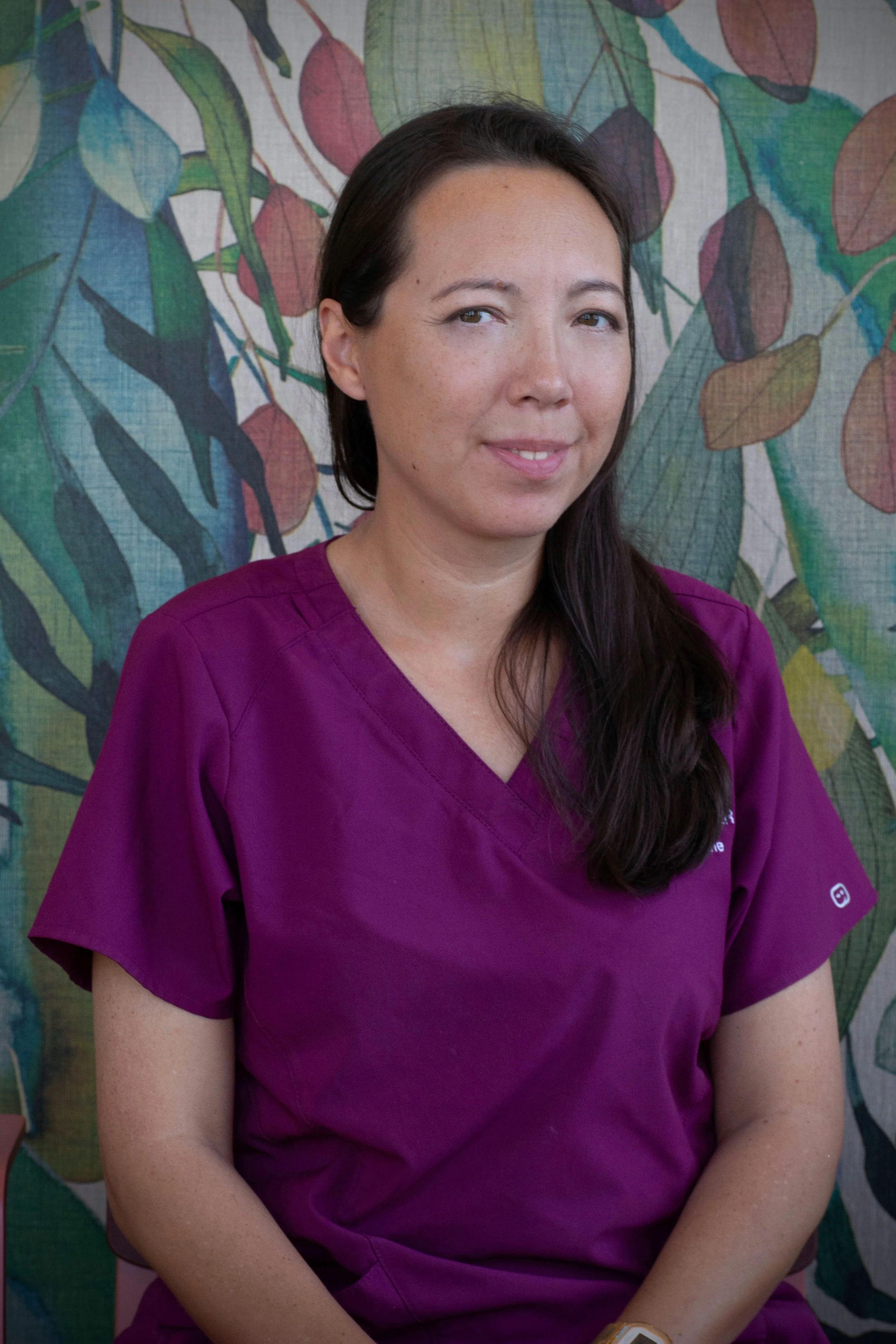 Kvinne med mørkt hår og svart t-skjorte som sitter ved et trebord, omgitt av grønne planter i et innendørs drivhus.