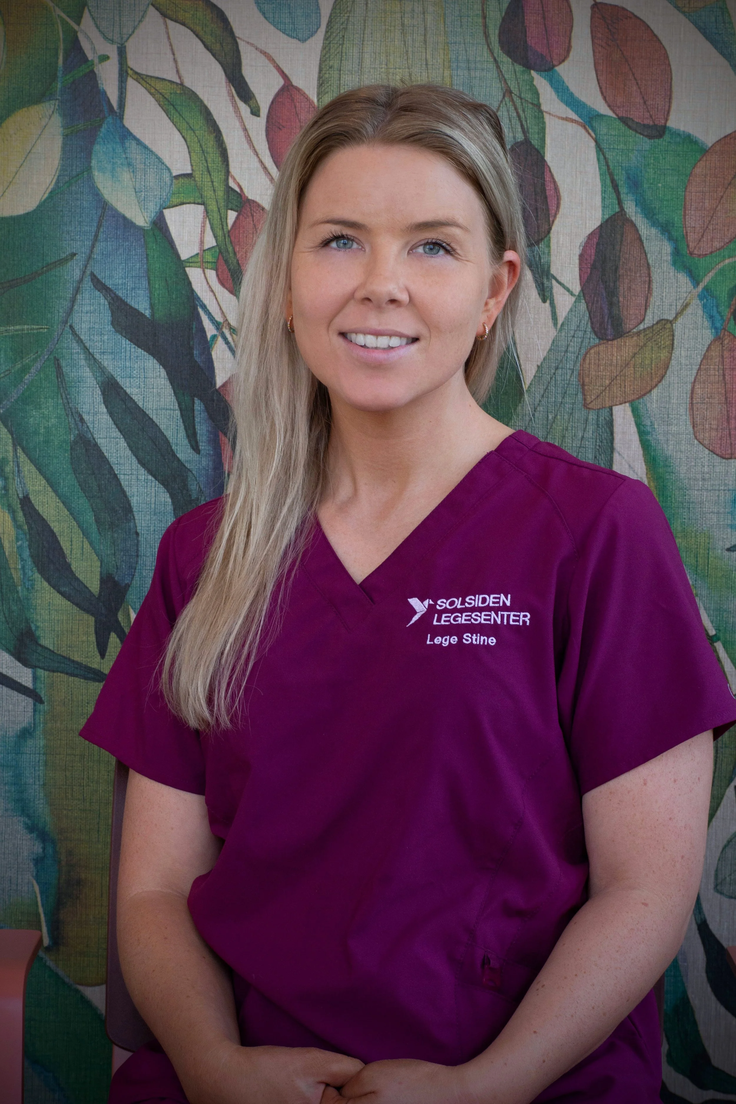 En smilende kvinne med blond hår, iført en svart genser, sitter ved et trebenk i en innendørs hage med grønne planter i bakgrunnen.