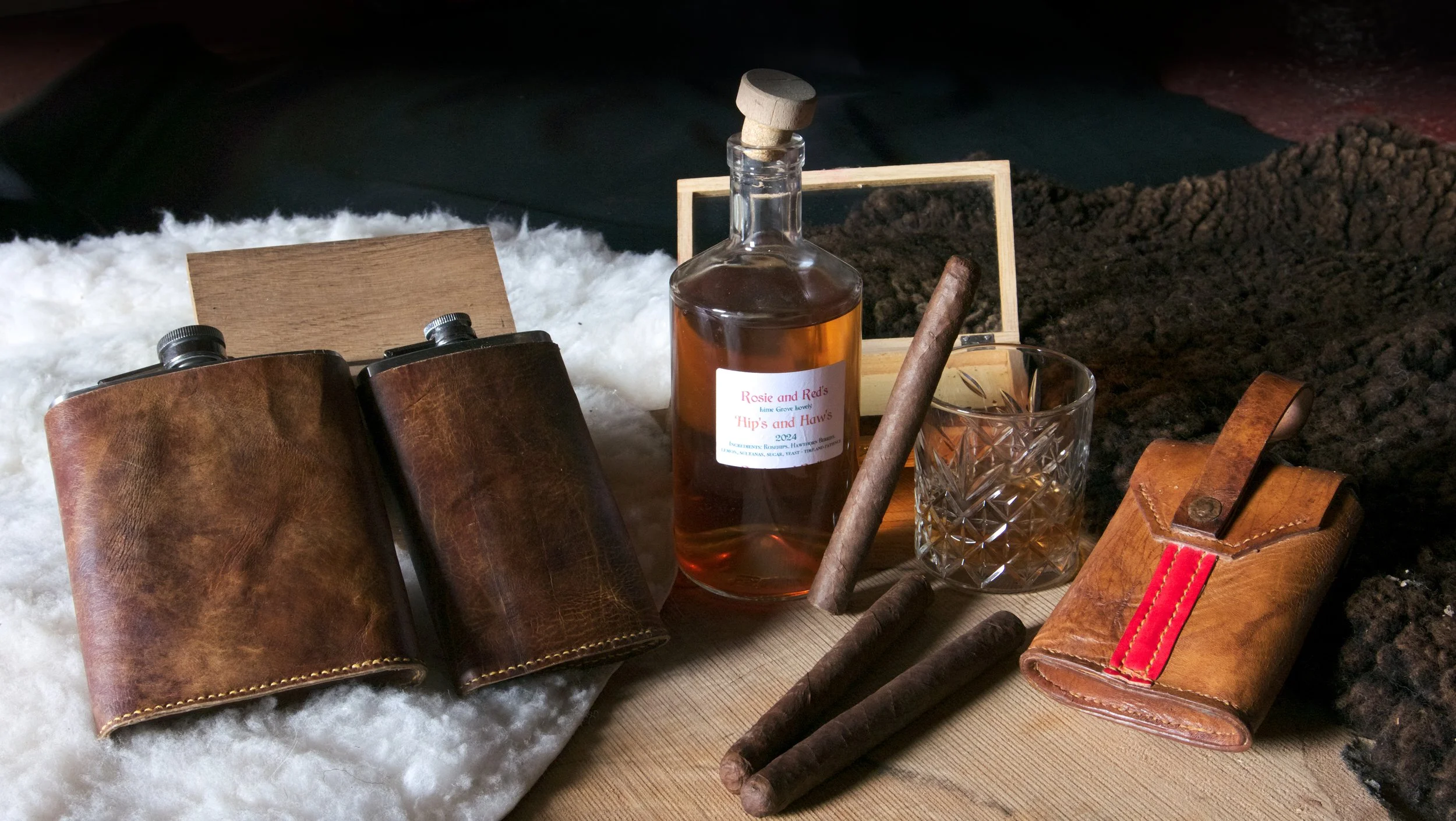 A collection of leather items, including two hand made leather flask holders, a small leather pouch, and a whiskey glass with a cigar and cigars on a wooden surface with a soft white and dark textured background.